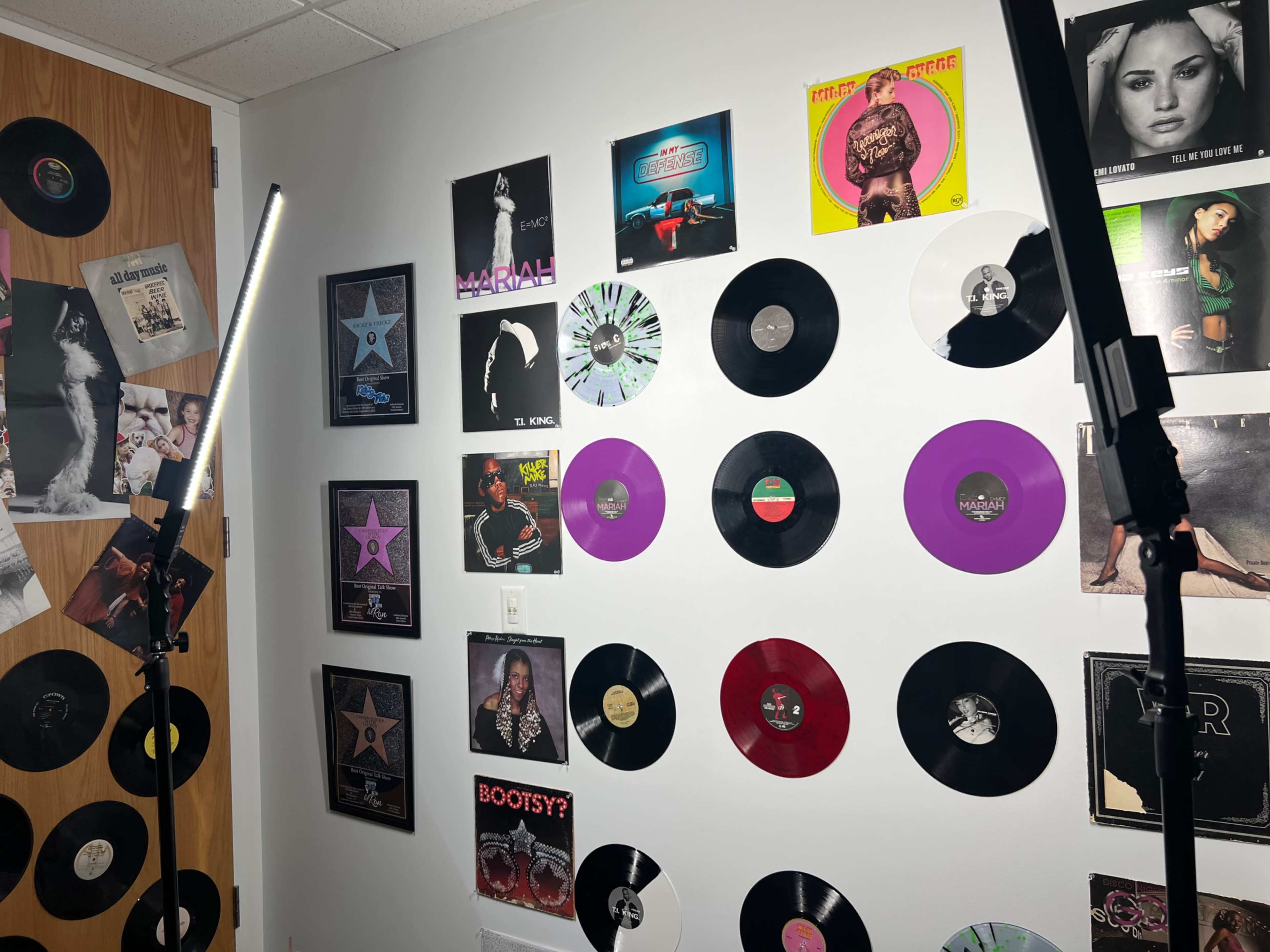 The image shows a room with a wall adorned with various vinyl records and framed music posters, along with two standing light fixtures.