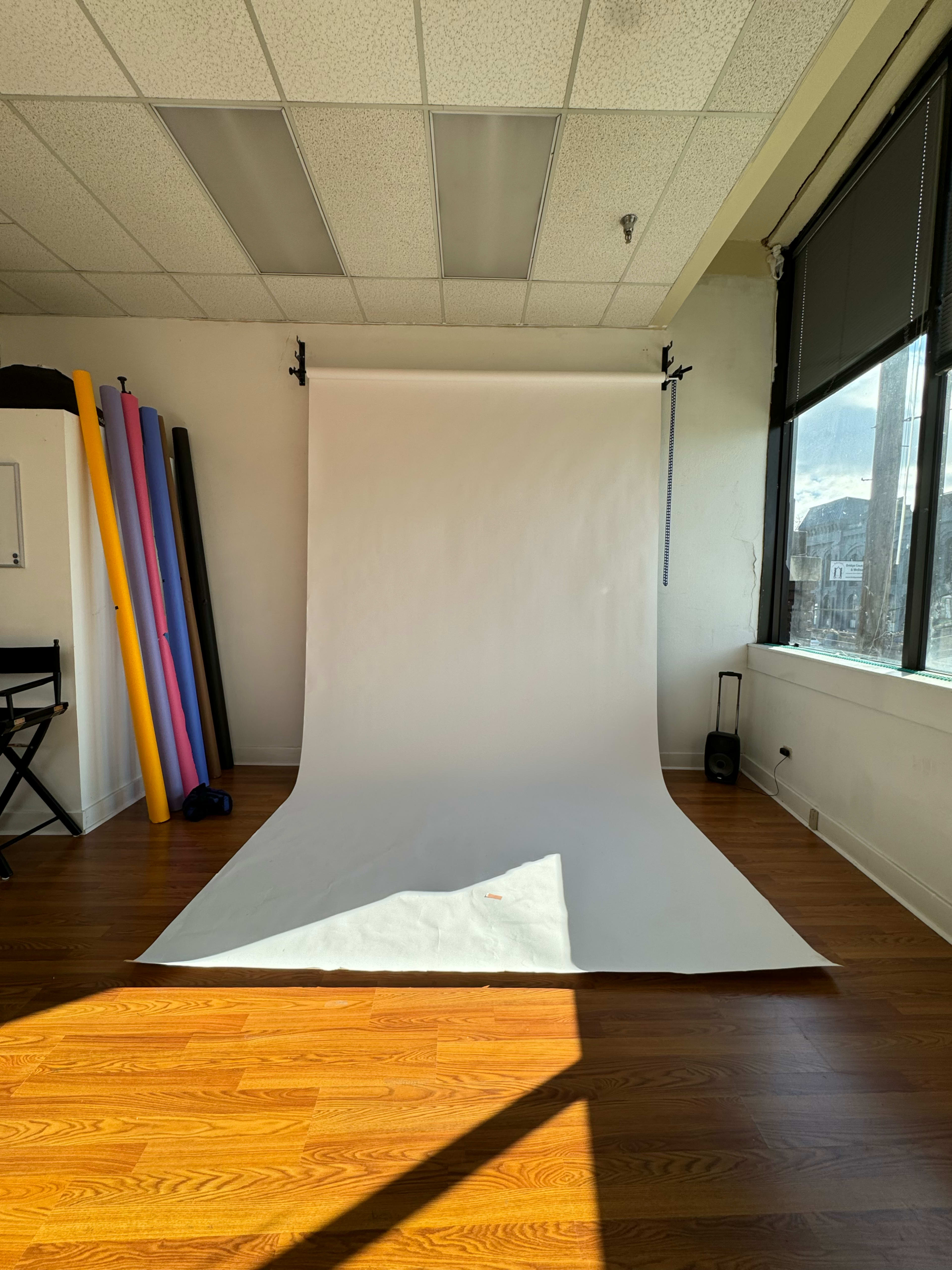 A photography studio features a plain white backdrop on a wooden floor, illuminated by natural light from a large window.