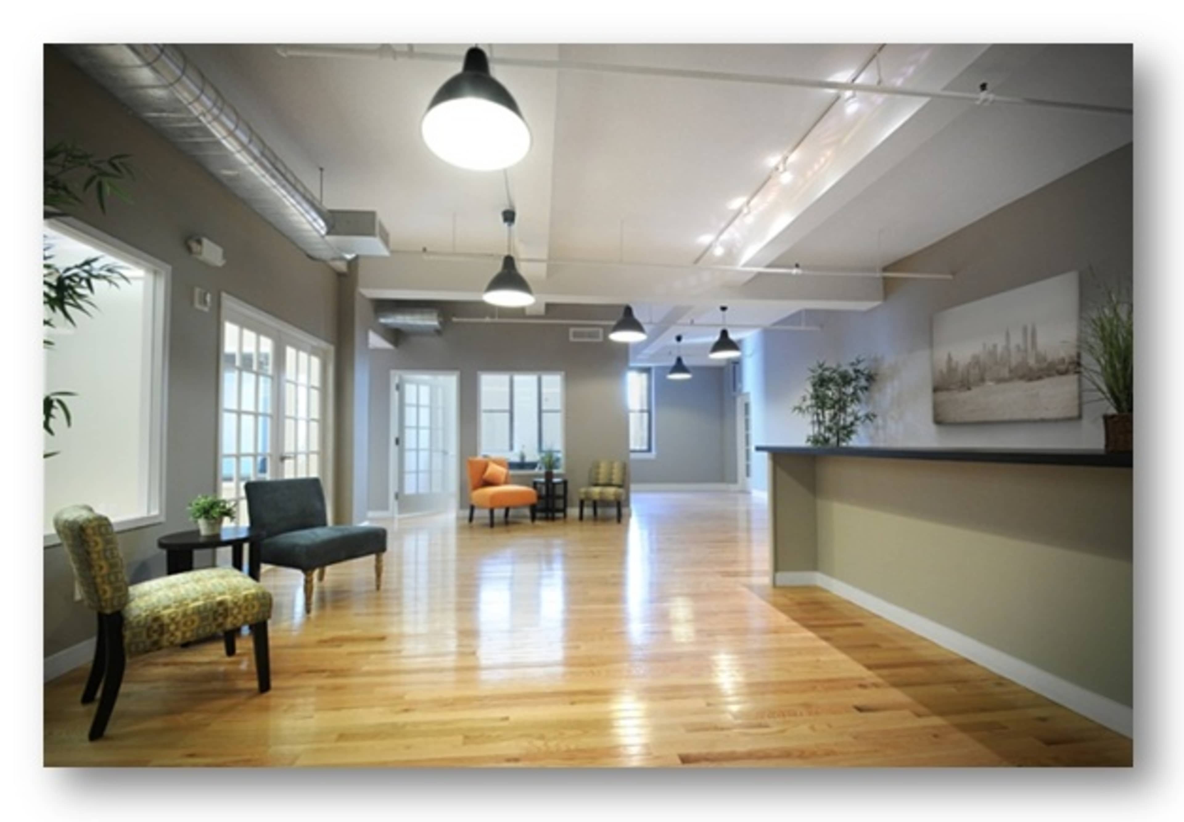 The image shows an empty, well-lit office space with wooden floors, contemporary furniture, and large windows.