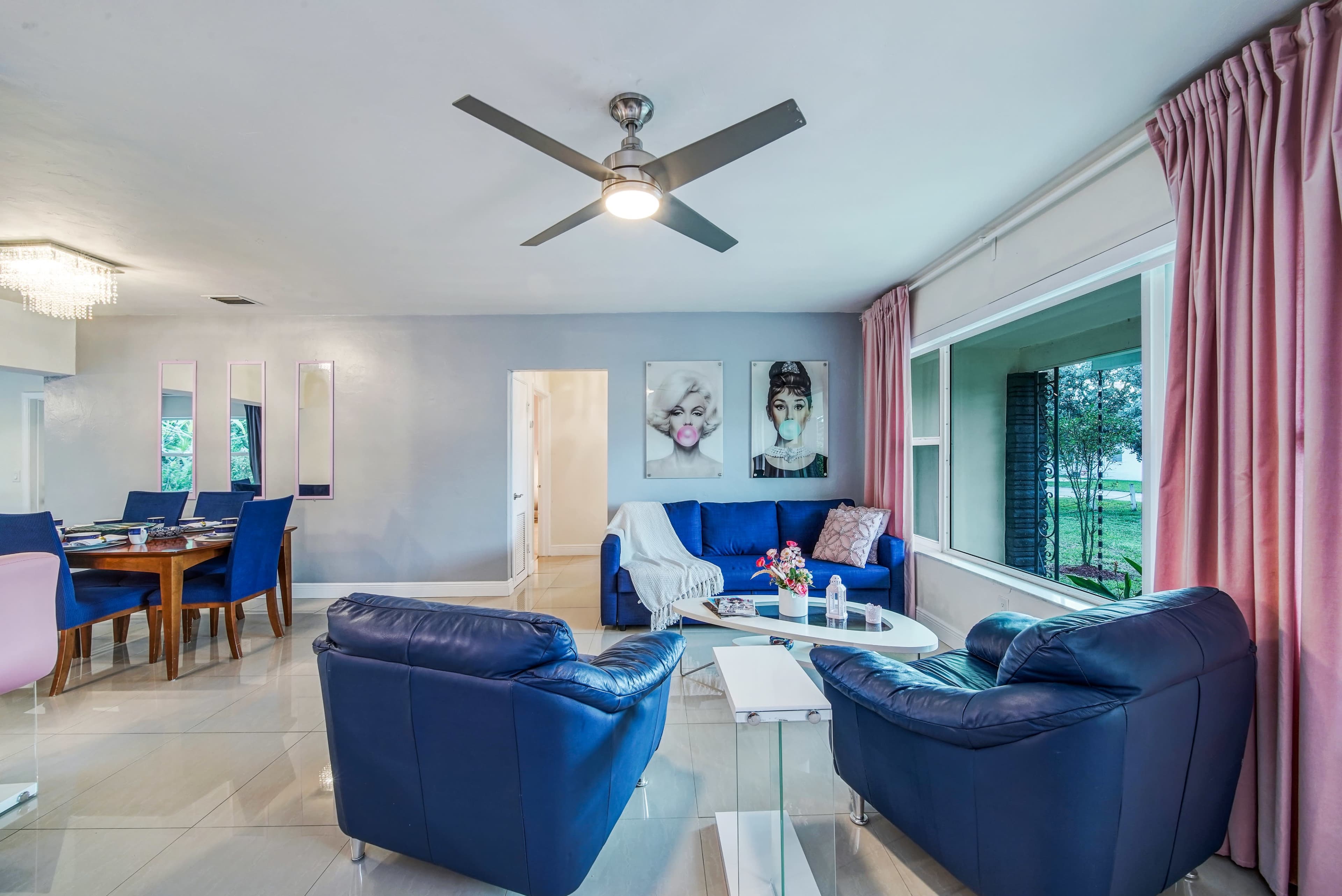 A modern living space features blue leather sofas, a glass coffee table, and a dining area with a wooden table, all set against light-colored walls and large windows.