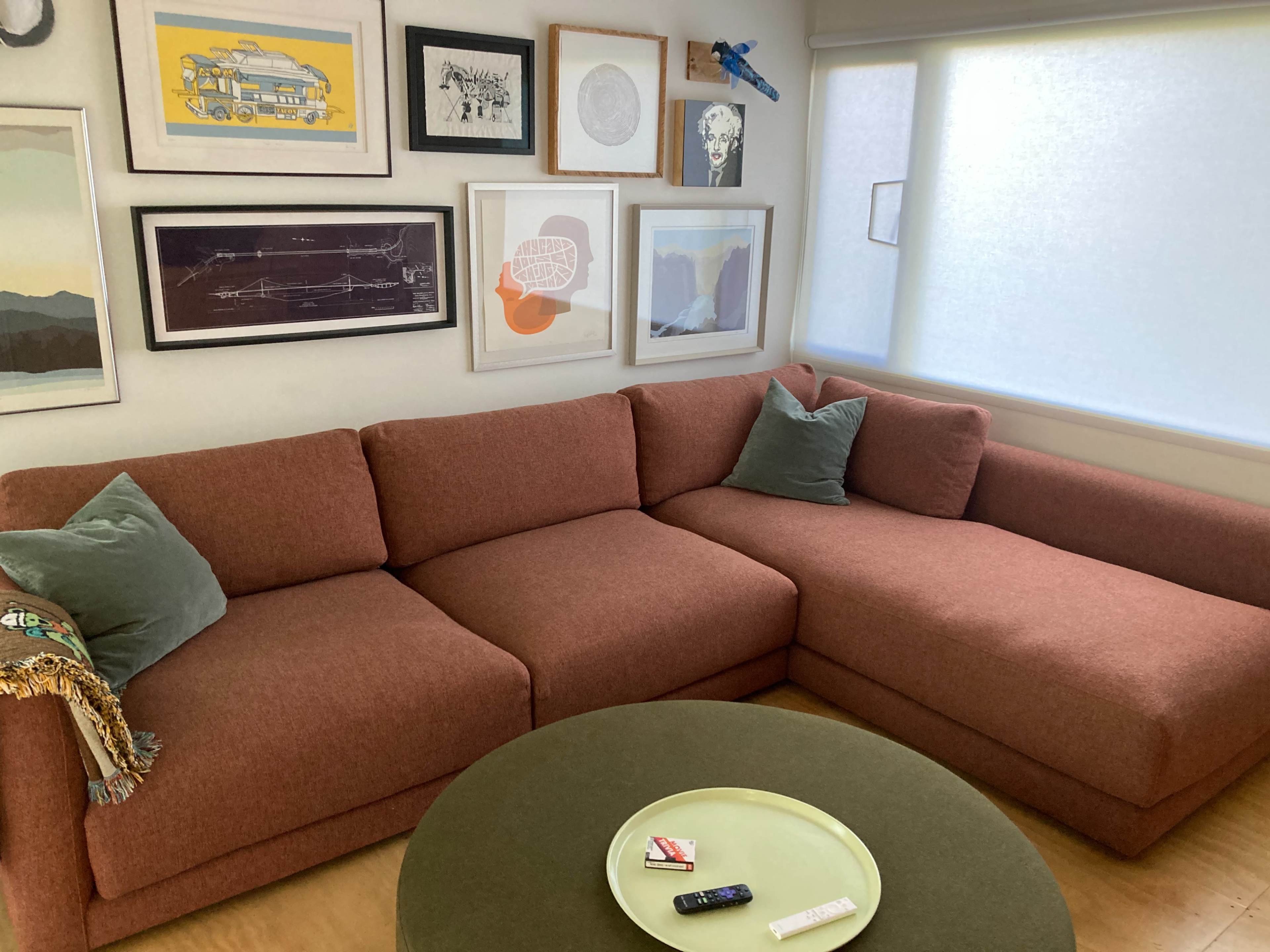 The image shows a modern living room featuring a large rust-colored sectional sofa with two green throw pillows, a round green coffee table, and a wall adorned with various framed art pieces.