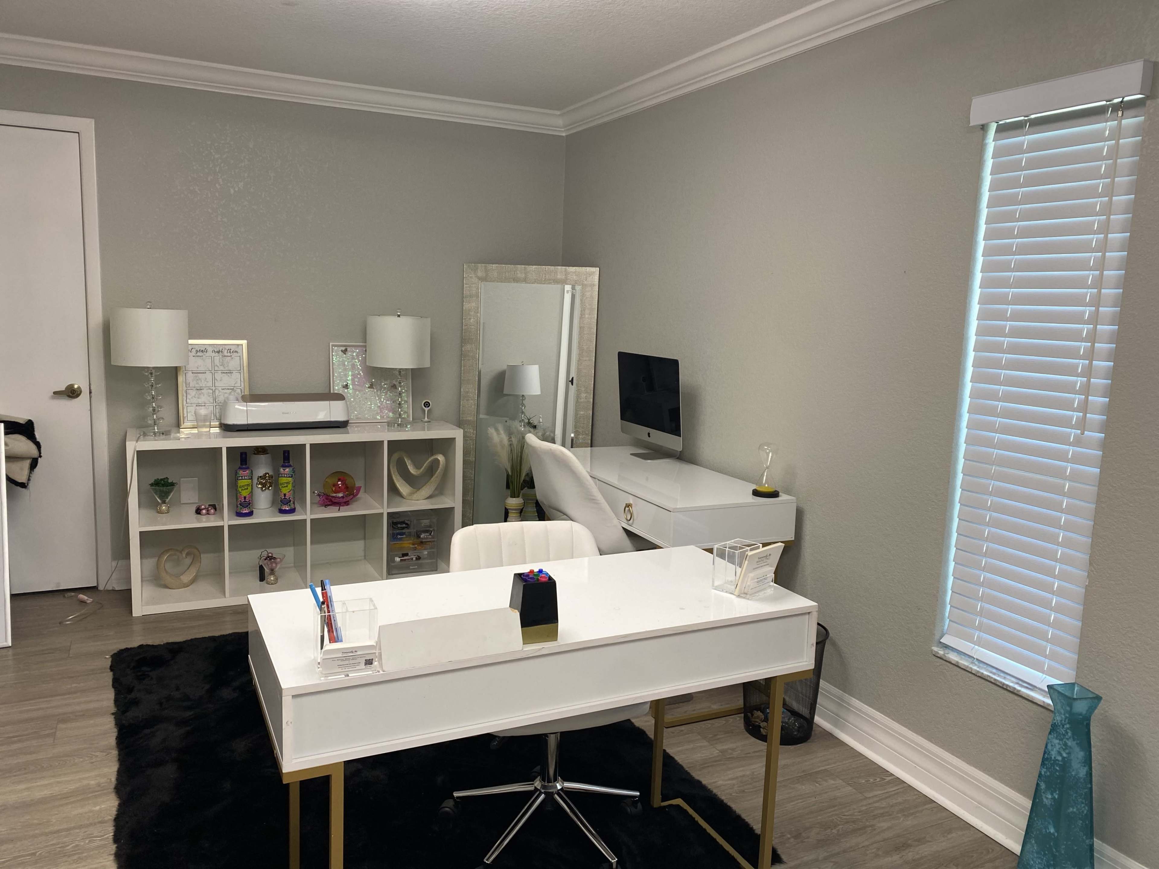 The image shows a modern office space featuring a white desk with a glass top, a chair, a shelving unit with decorative items, and a large mirror along with a computer setup against the wall.