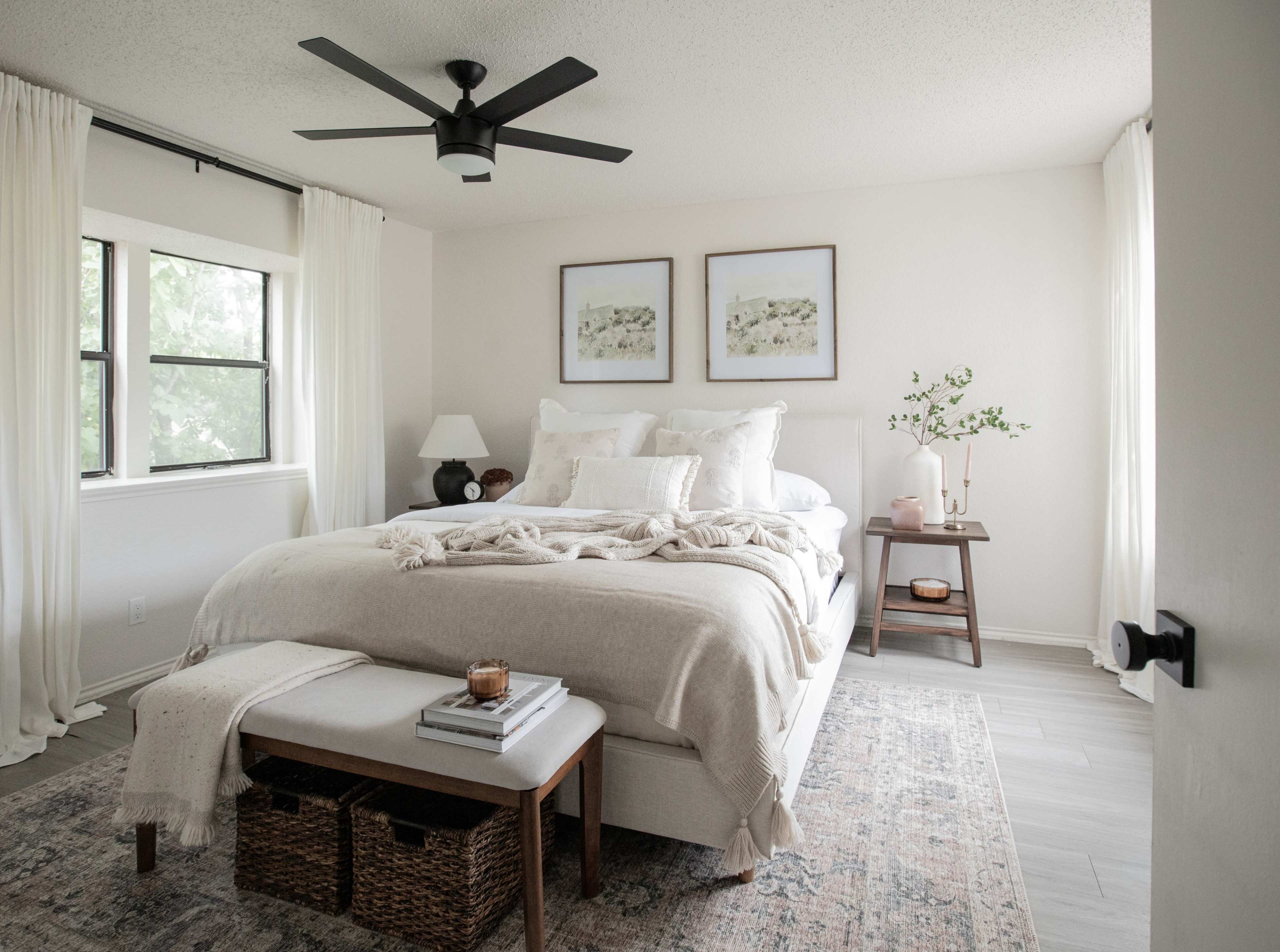 A well-lit bedroom features a large bed with white bedding, a bedside table with a lamp, and framed artwork on the walls.