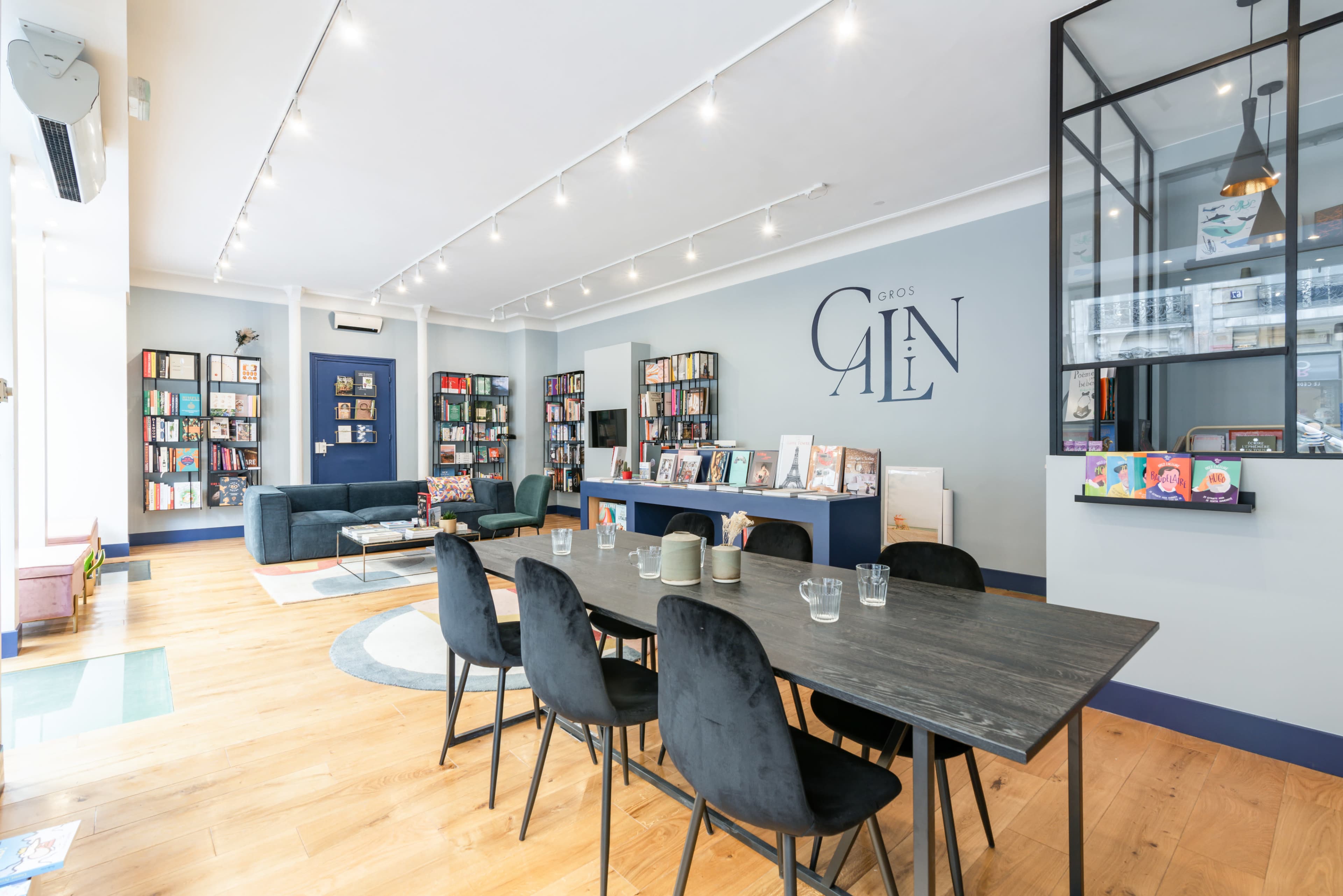 A modern, well-lit community space with bookshelves lining the walls, a large wooden dining table surrounded by black chairs, and comfortable seating areas.