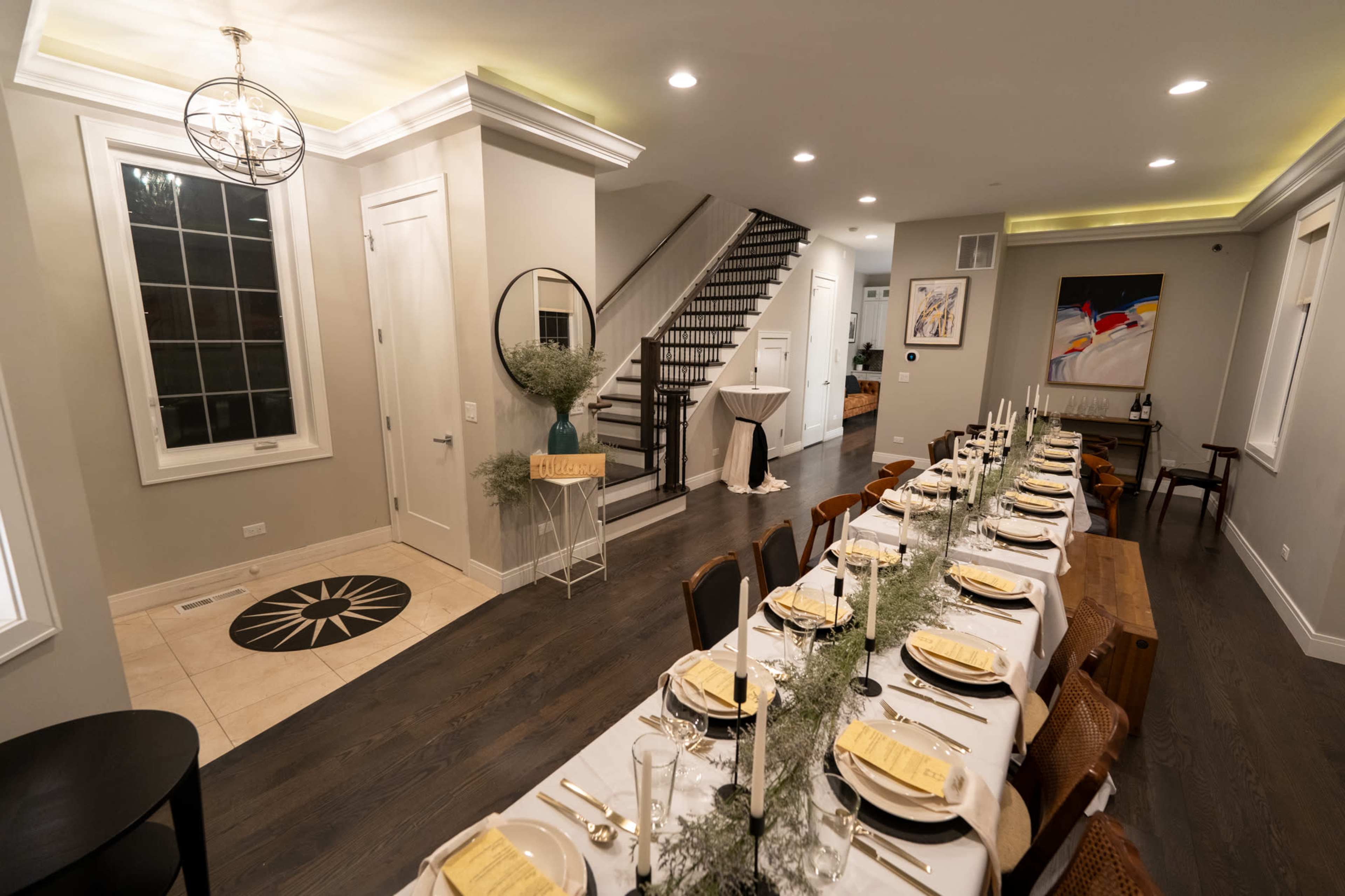 A long dining table is set for a meal in a modern, well-lit room that features a staircase leading to another level.