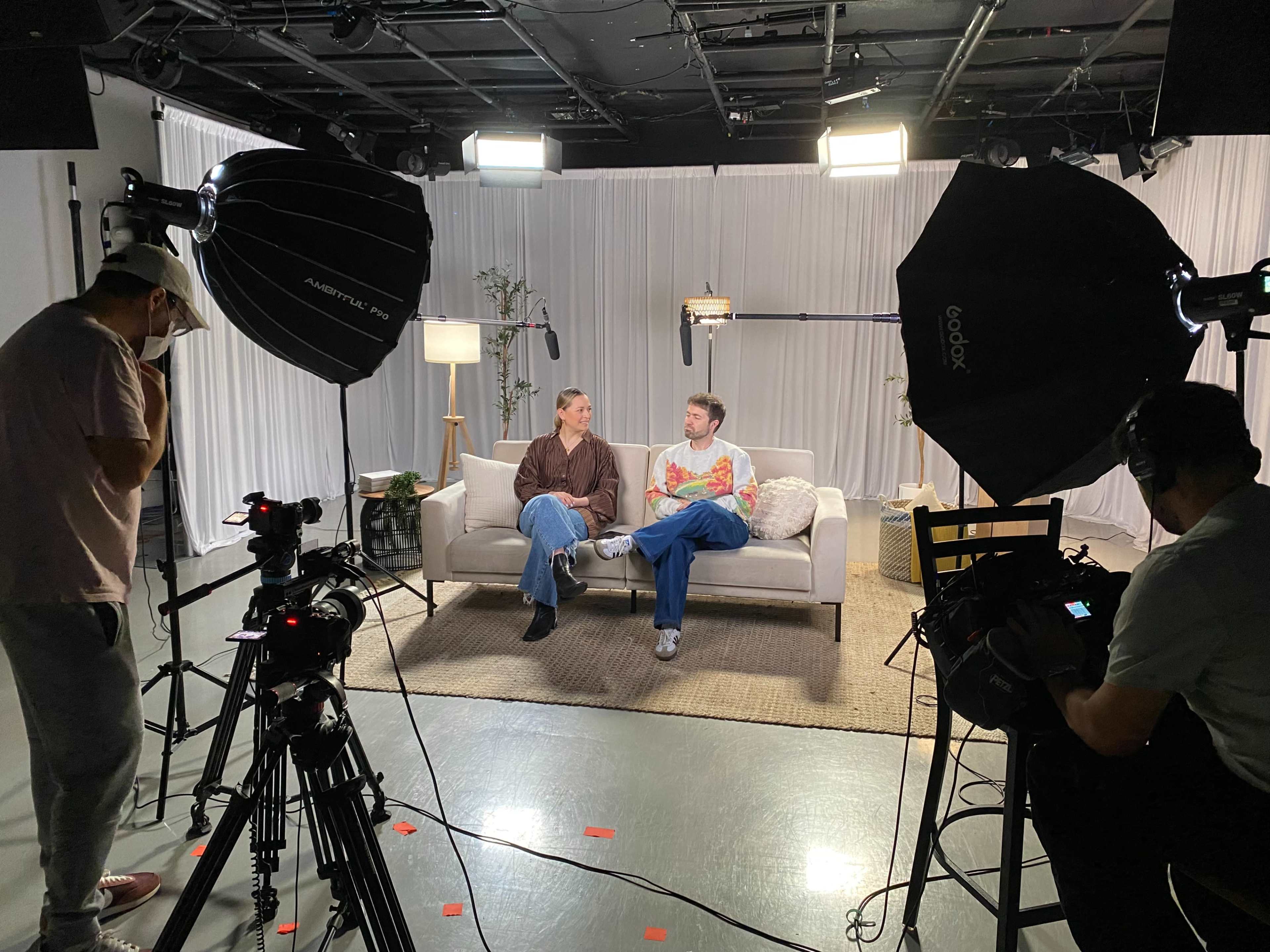A television studio set features two individuals seated on a couch, surrounded by cameras and lighting equipment.