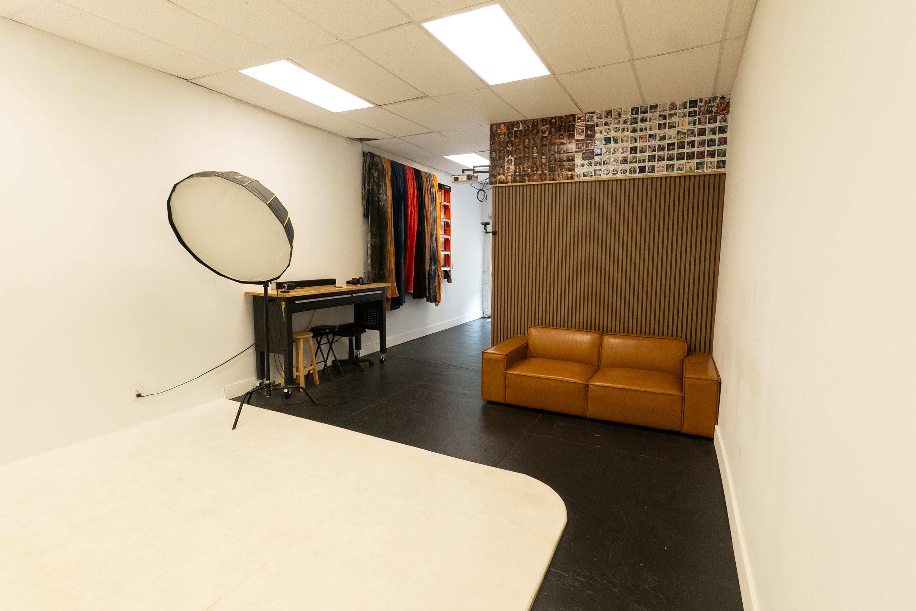 The image shows a photography studio featuring a light reflector, a small table with equipment, a brown sofa, and wall décor comprised of photographs.