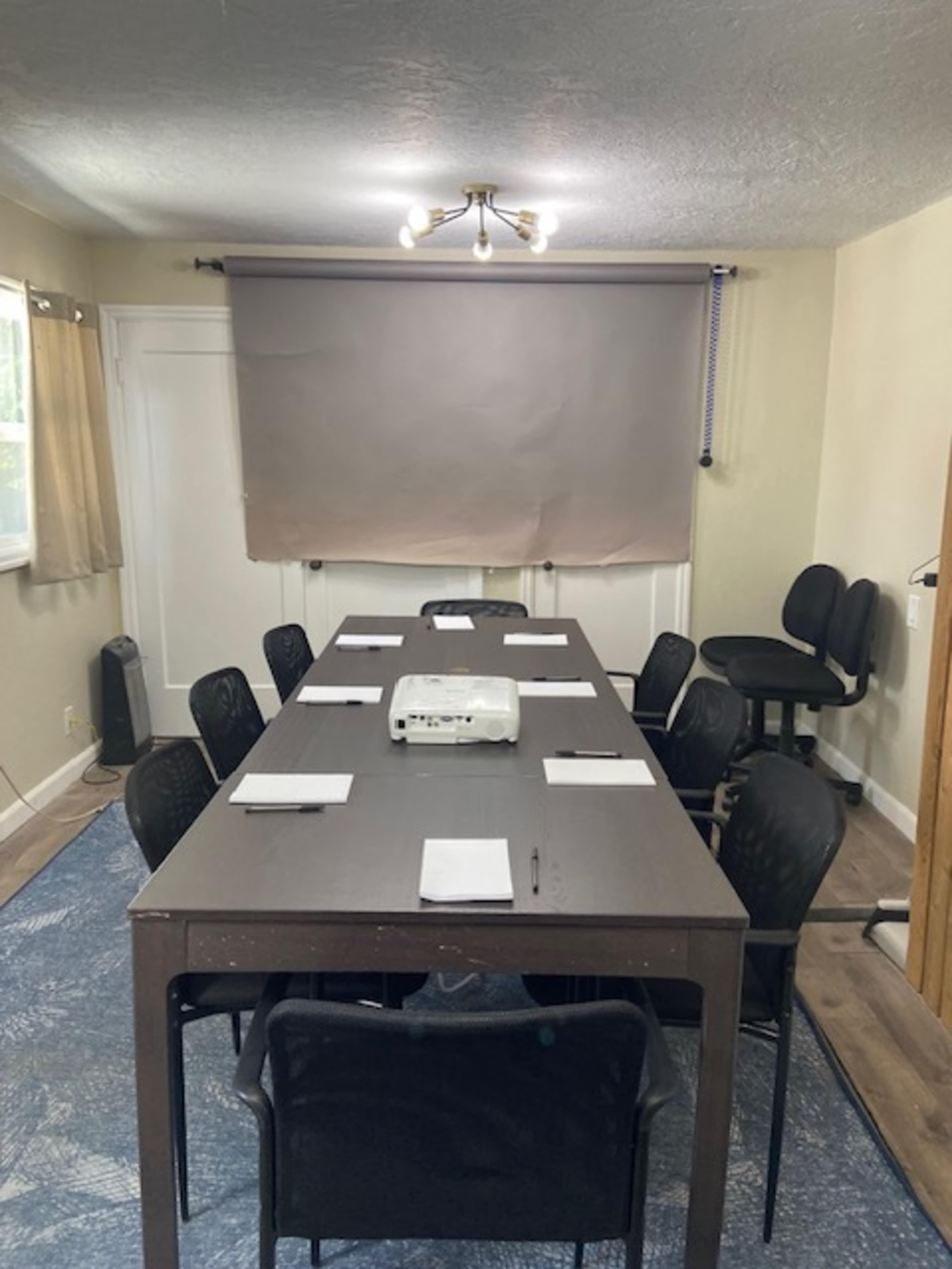 A long table with chairs is arranged in a well-lit room, featuring a projection screen and a projector positioned on the table.