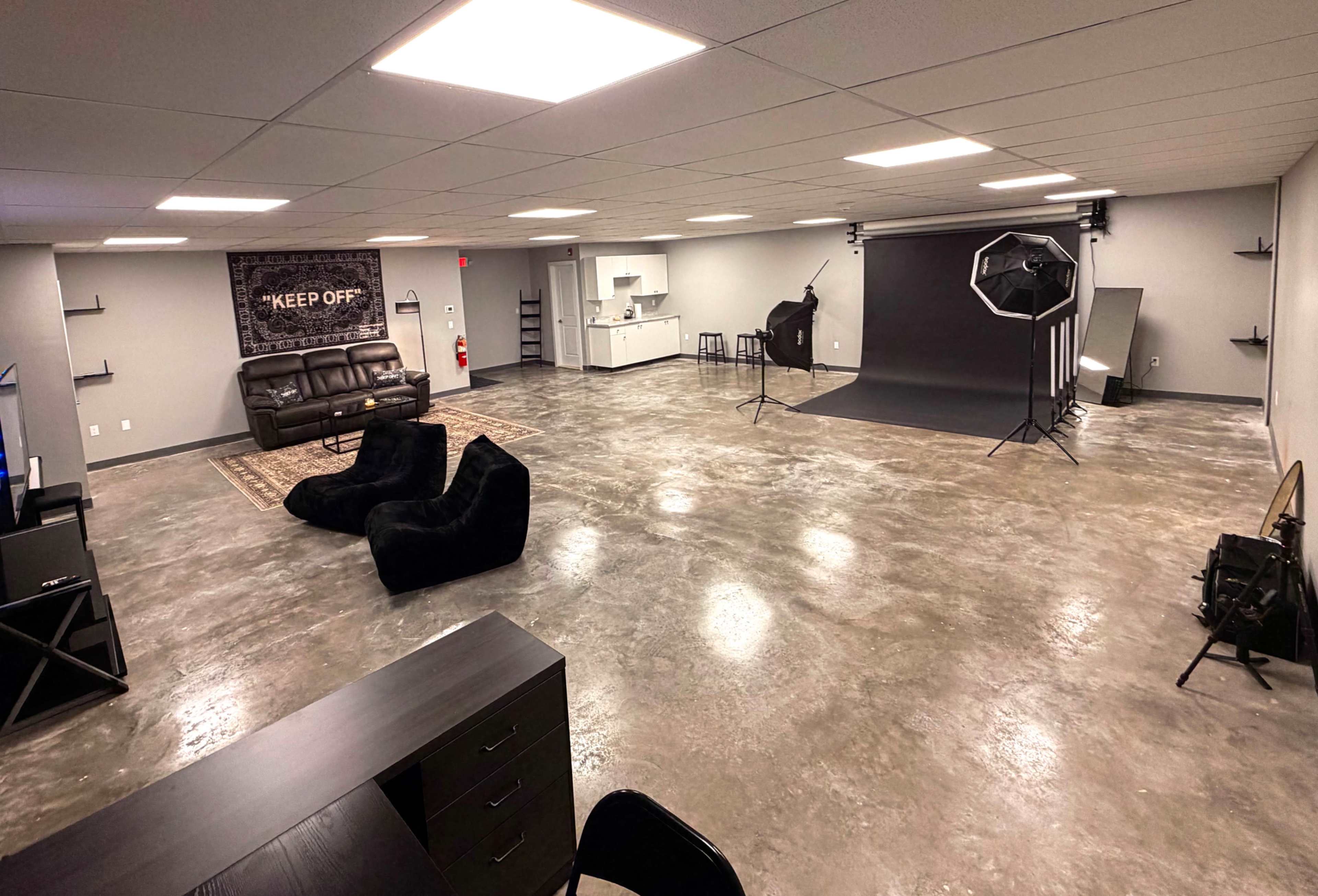 The image shows a spacious studio interior with polished concrete floors, a couch, bean bag chairs, a kitchenette area, and photography equipment set up in one corner.