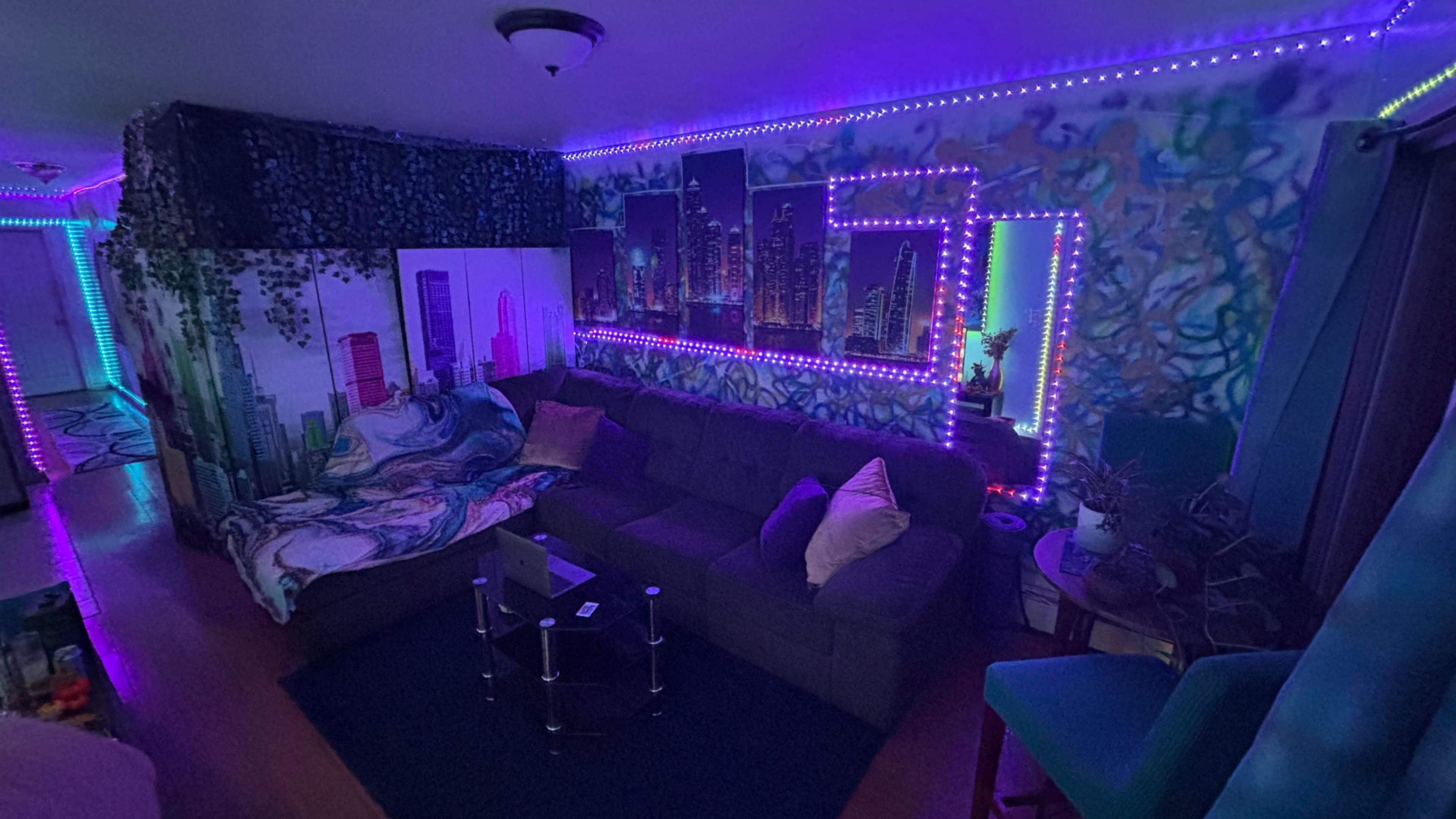 The image shows a cozy living room featuring a large gray couch, colorful wall art, and vibrant LED lights illuminating the space.