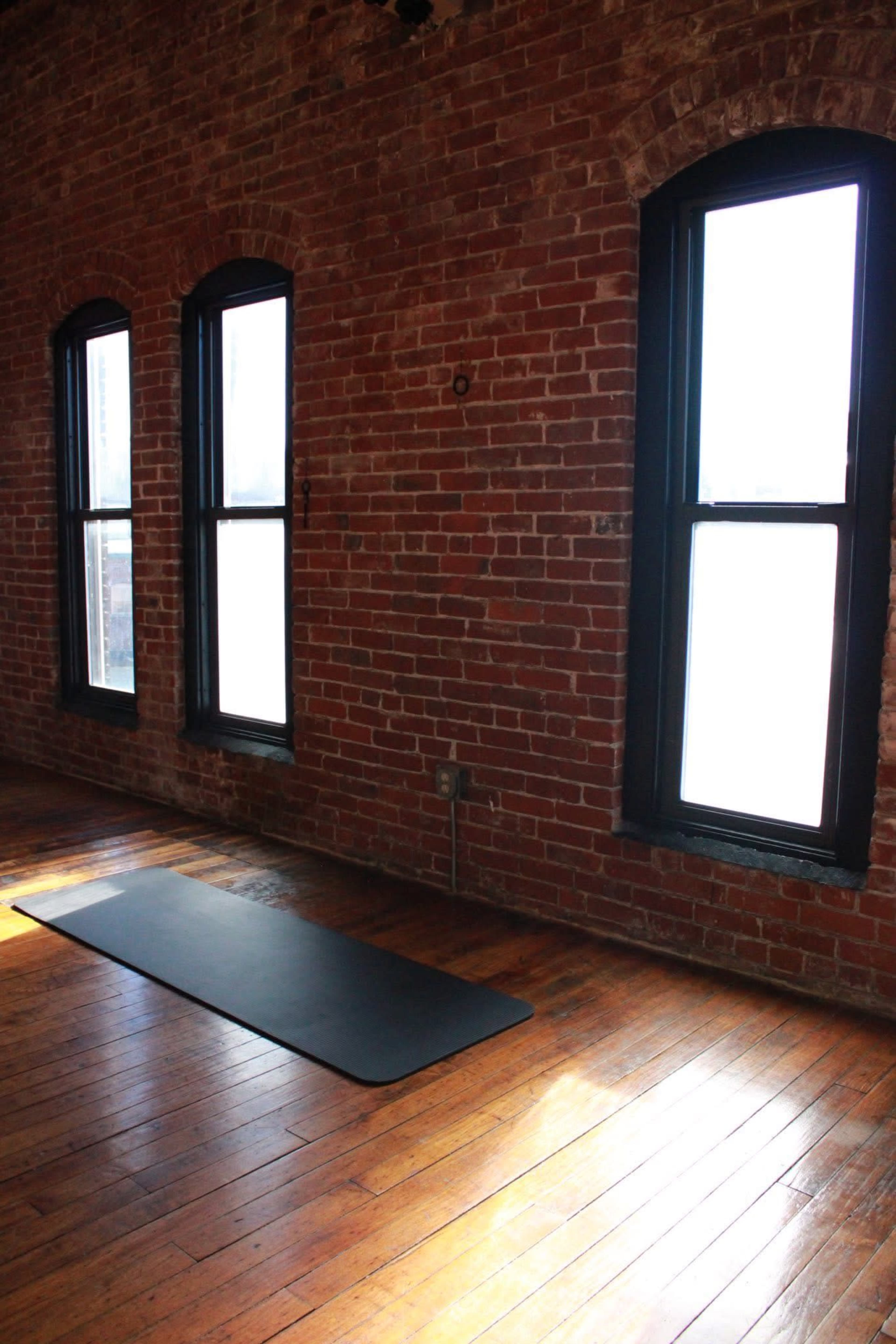 The image shows a spacious room with exposed brick walls, three windows, and a yoga mat placed on a polished wooden floor.