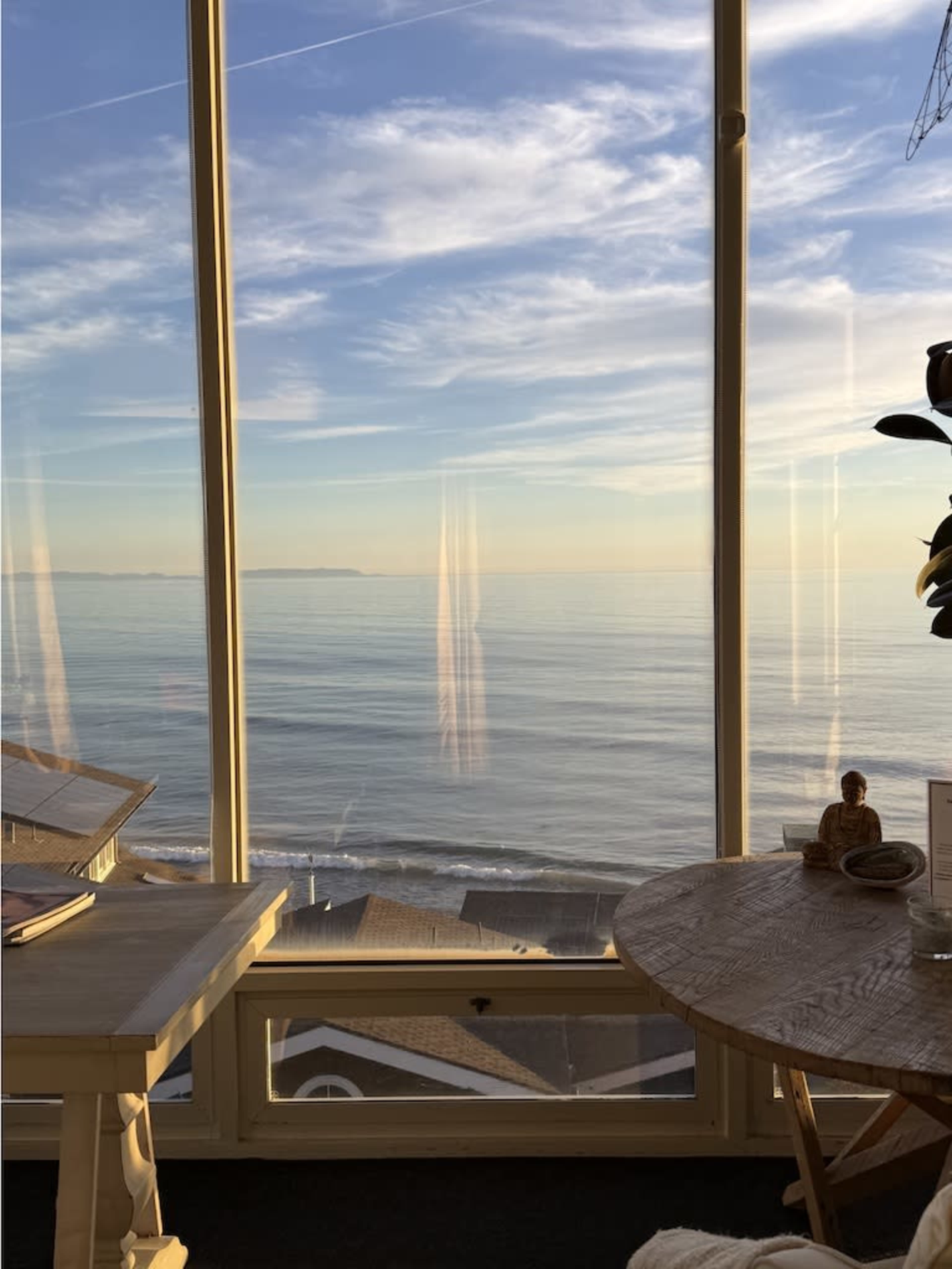 A large window offers a view of calm ocean waters under a clear sky at sunset, with a table and decor in the foreground.