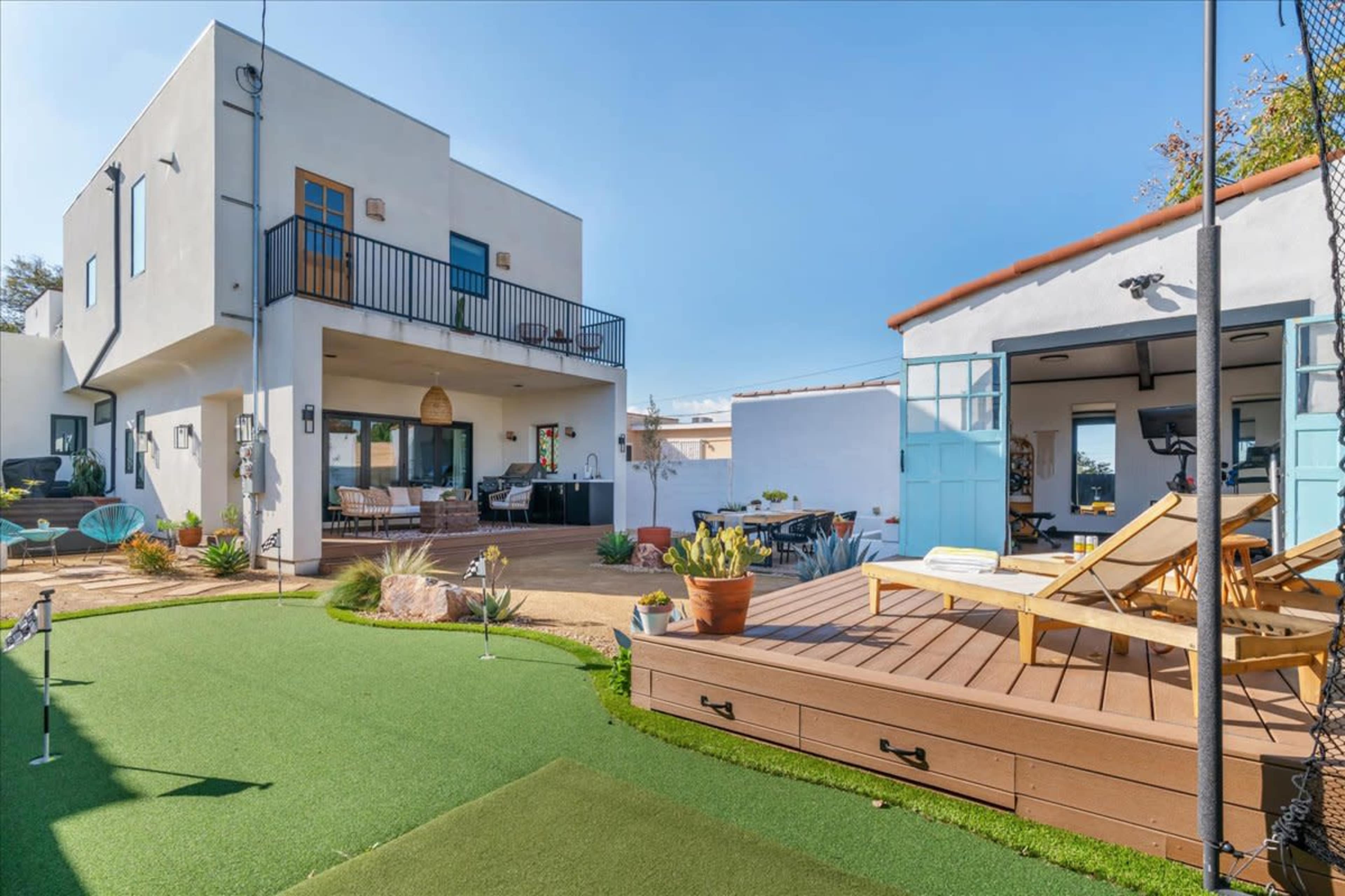 The image shows a modern two-story house with a large patio area, outdoor seating, and a small putting green in the foreground.