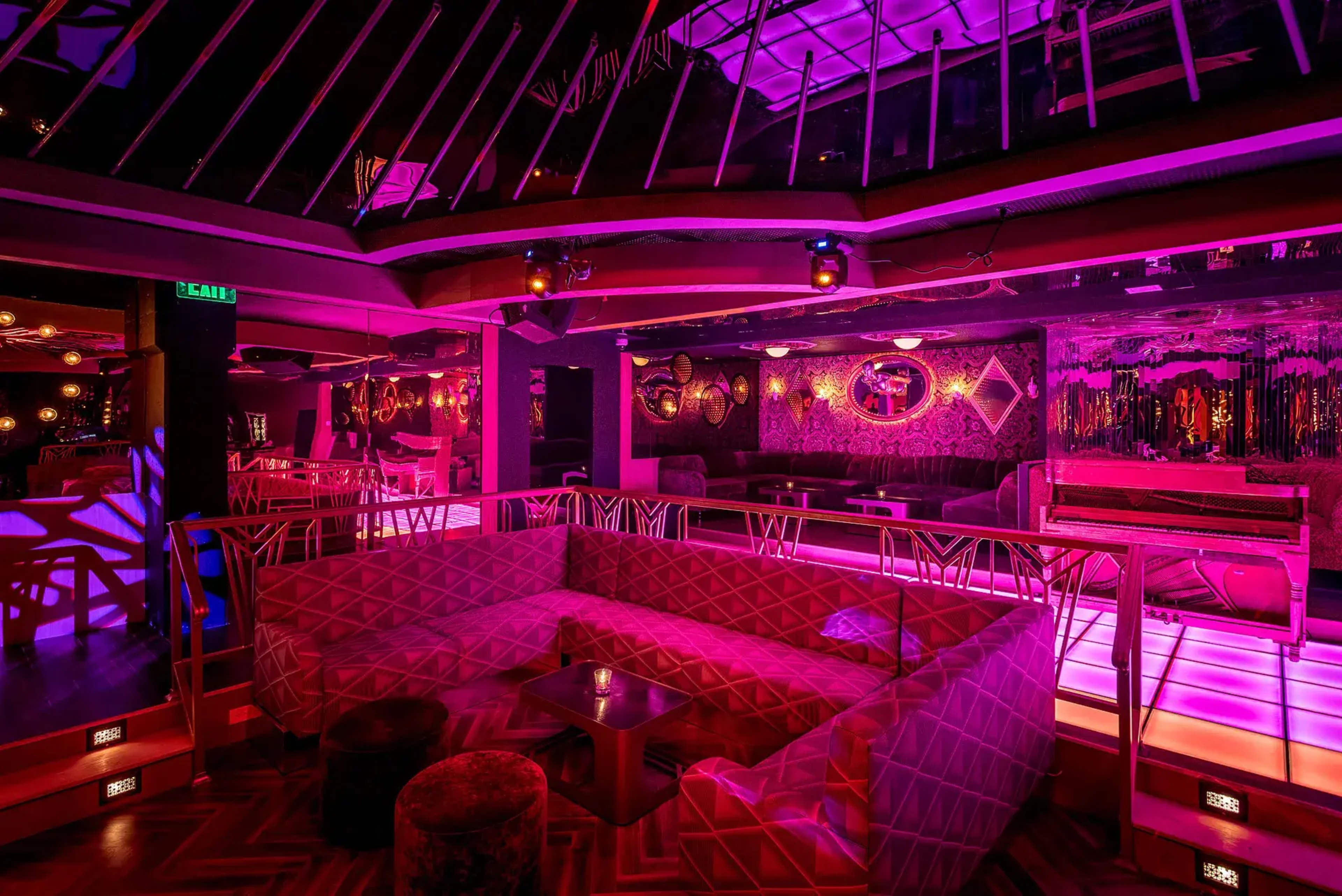 The image shows a dimly lit lounge area with purple lighting, featuring a large geometric sofa and scattered seating arrangements.