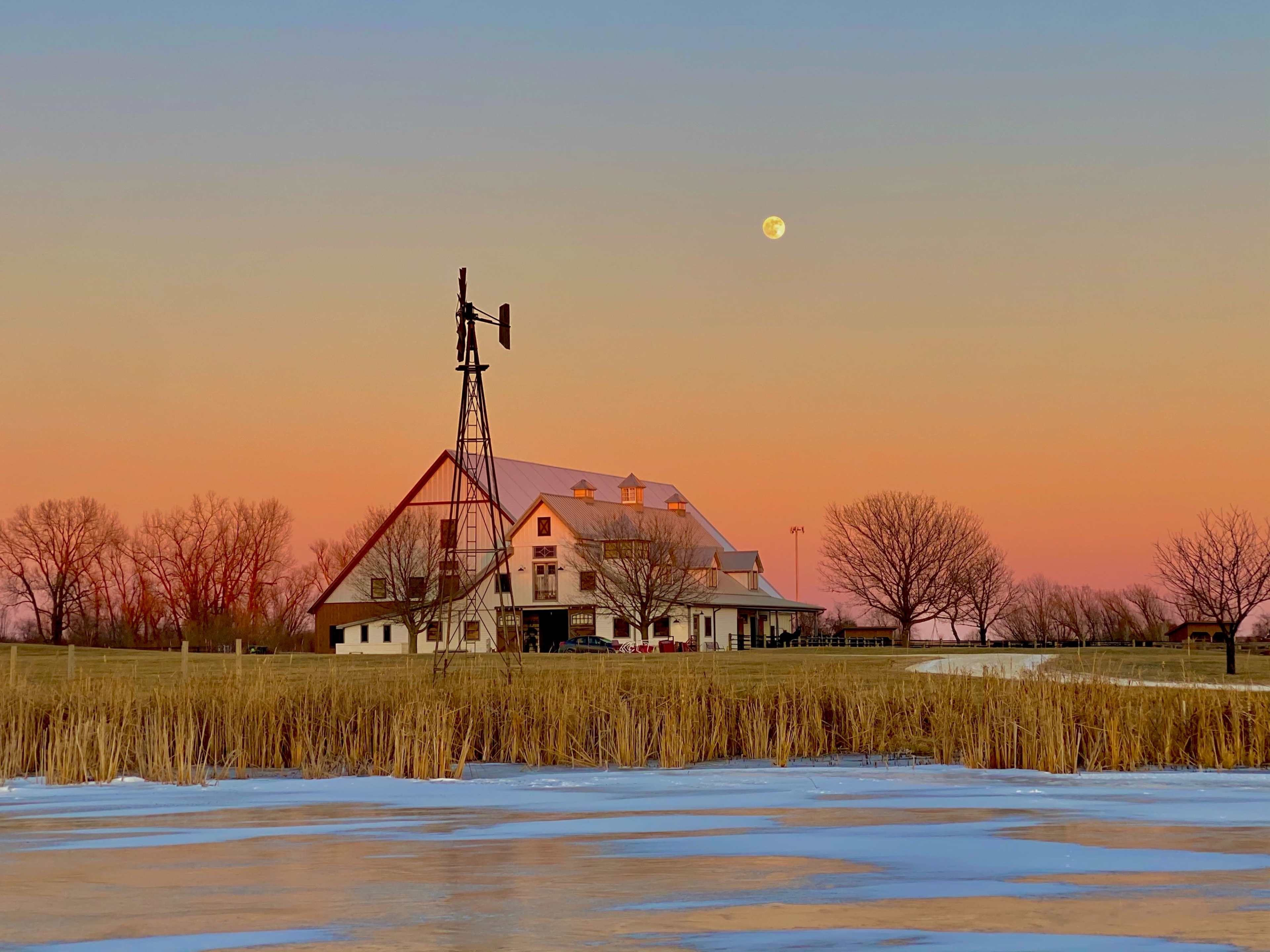 A white barn with a windmill is situated near a frozen pond, with a clear sky showcasing a rising moon at sunset.