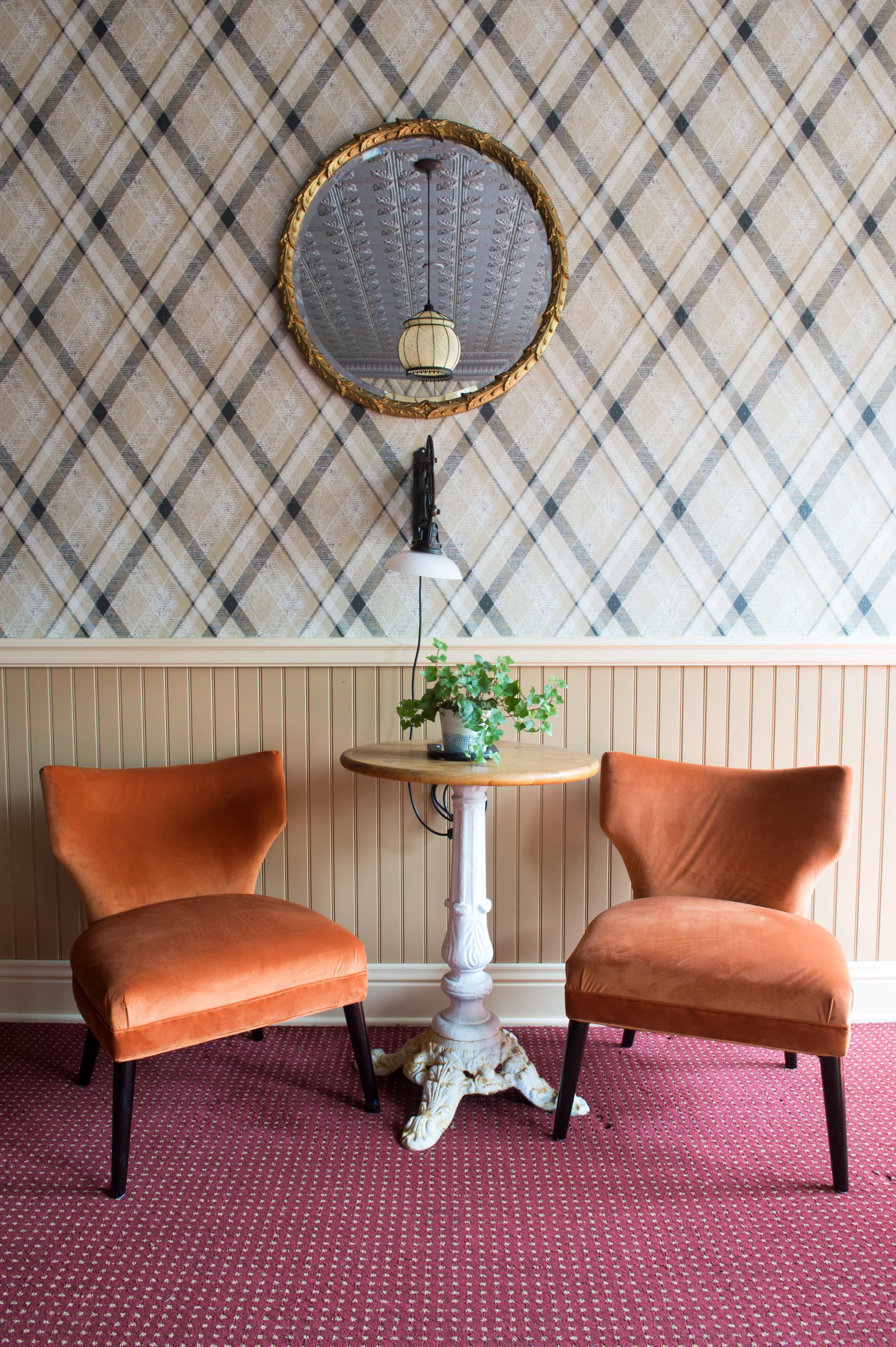 Two orange velvet chairs positioned around a small round table with a potted plant, set against a plaid wallpaper background.