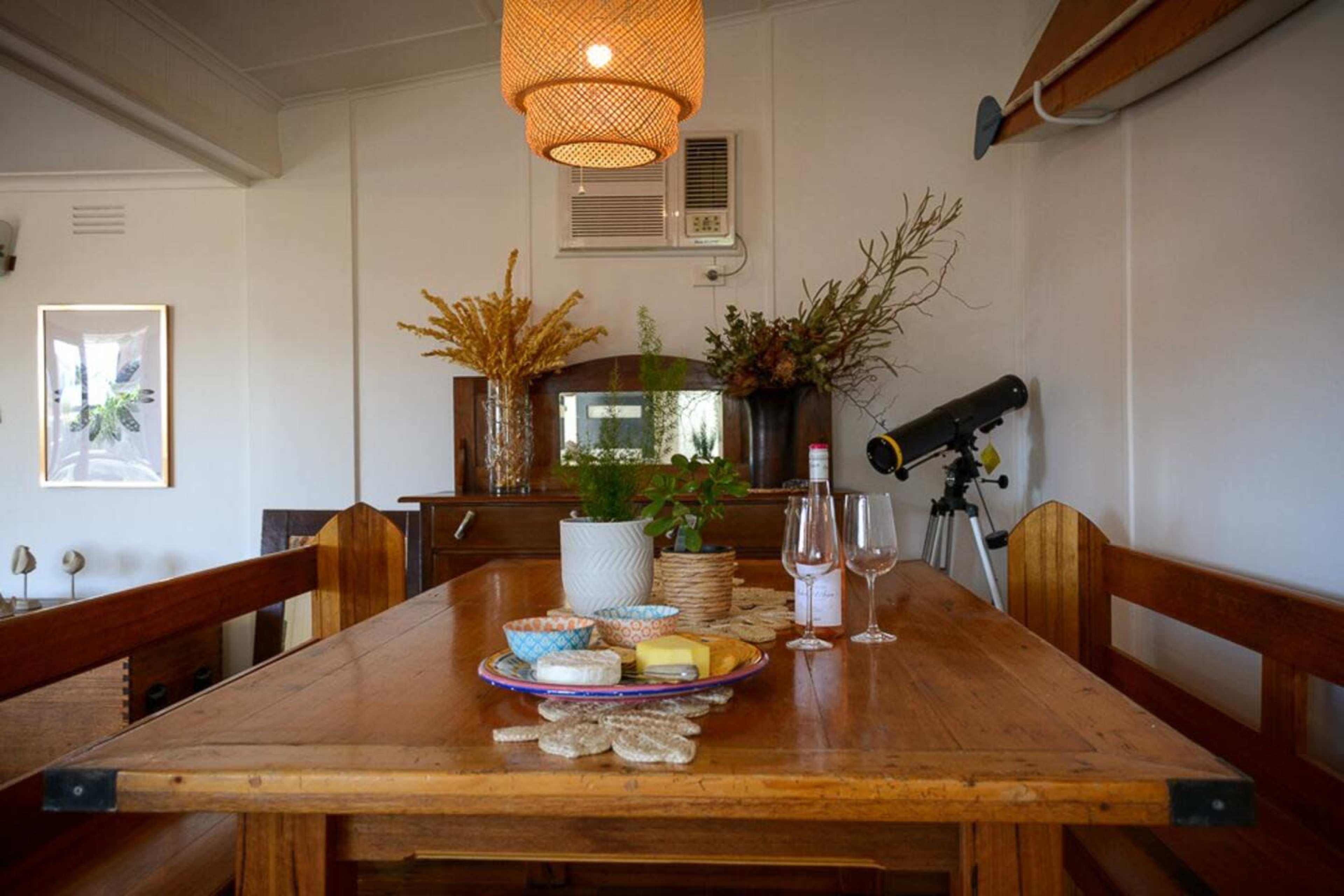 A wooden dining table is set with a plate of cheese, crackers, and glasses beside a telescope, under a woven light fixture in a decorated room.