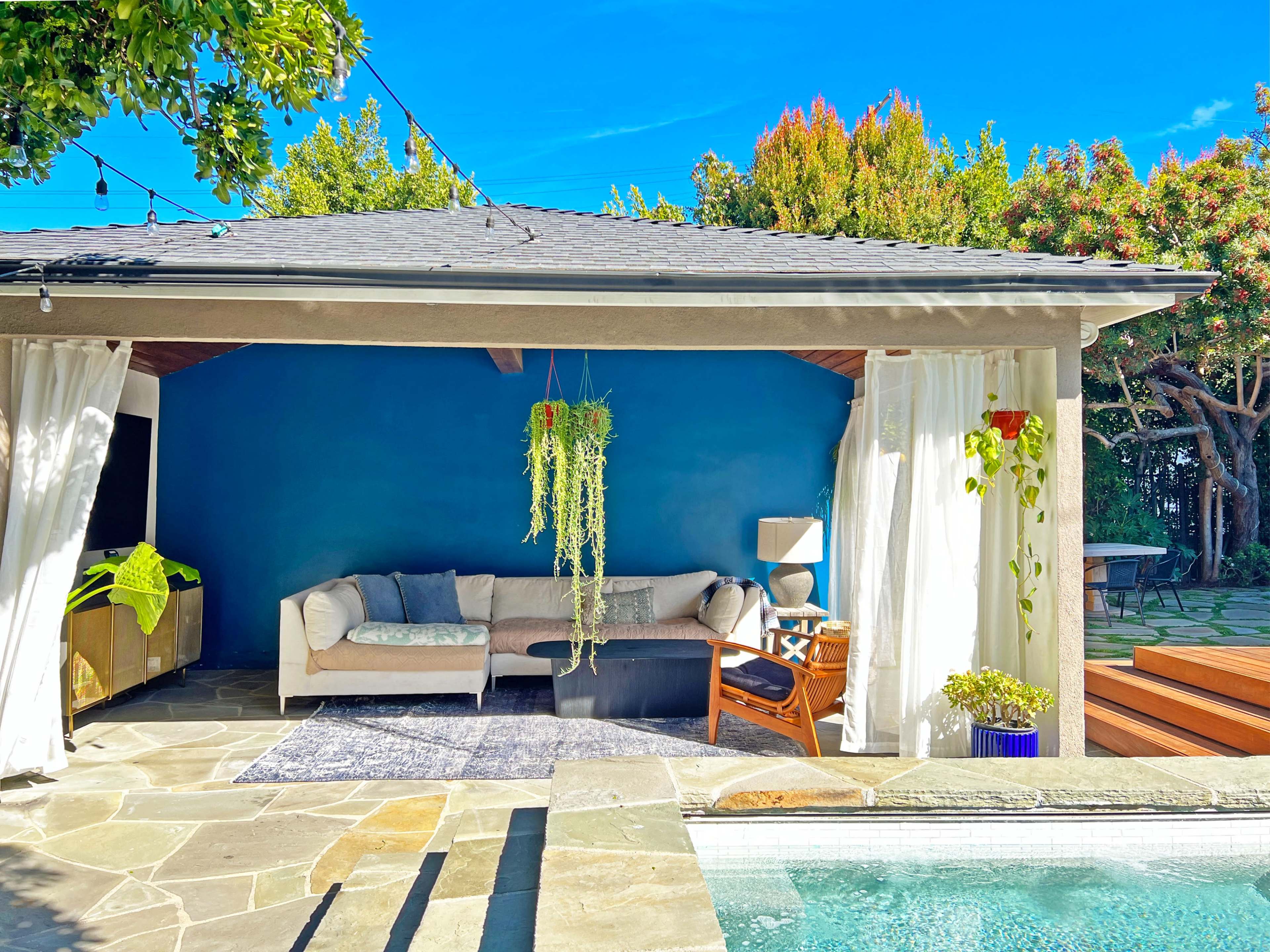 A covered outdoor living space features a light-colored sofa, a rug, hanging plants, and a backdrop of blue walls.