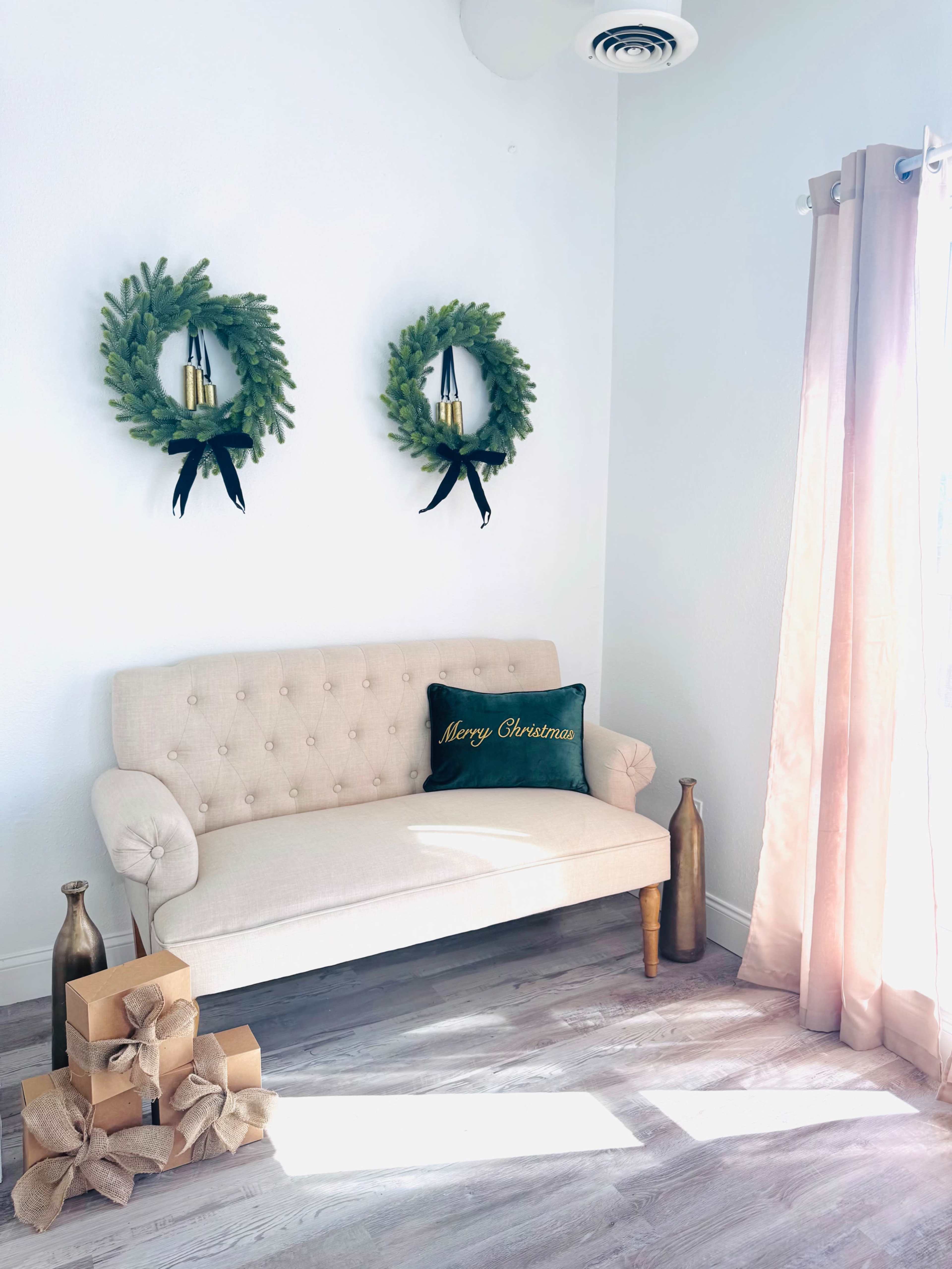 A light-colored sofa with a decorative Christmas pillow sits against a wall adorned with two green wreaths, while gift boxes are placed on the floor beside the sofa.