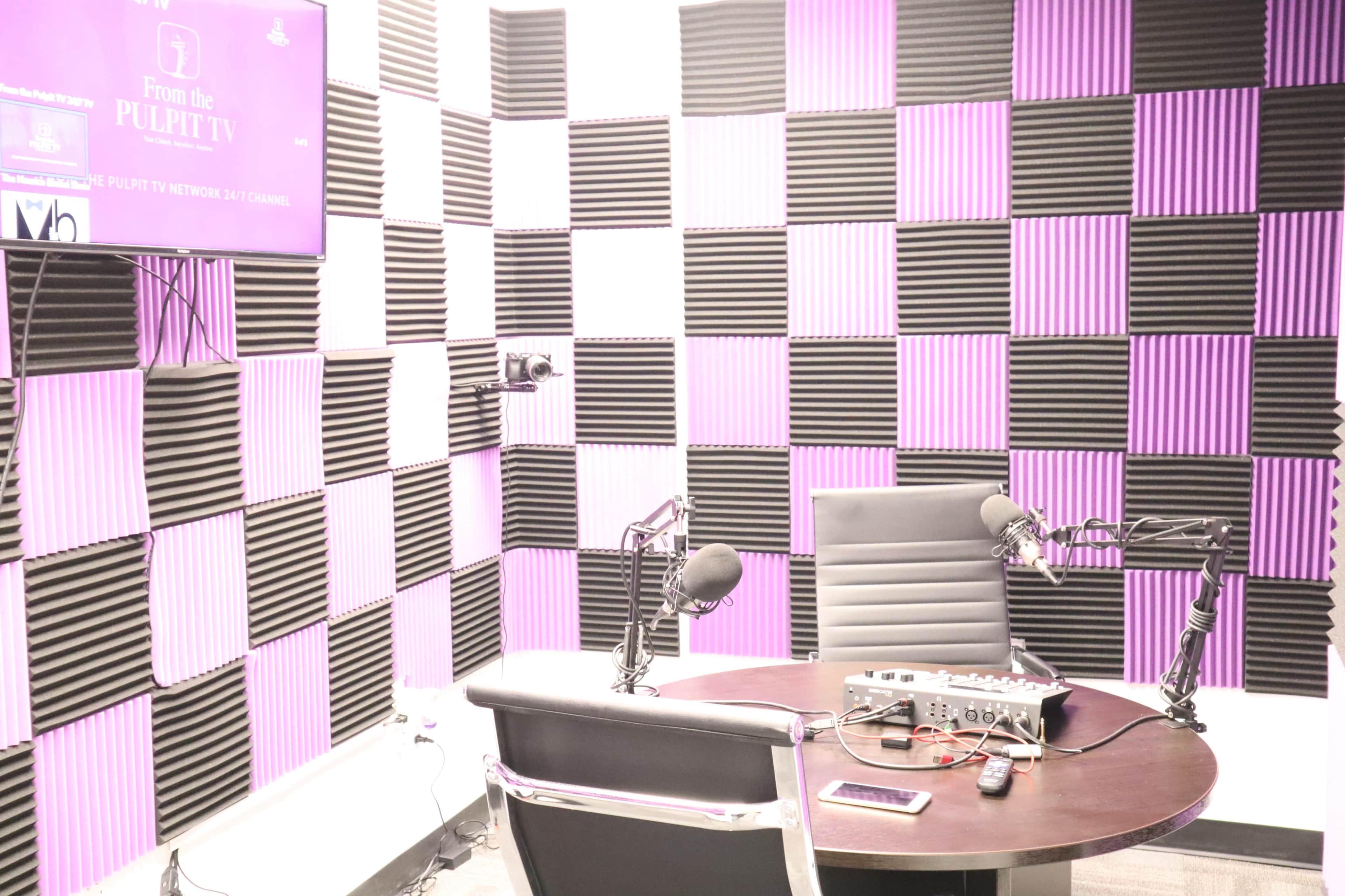 A recording studio with black and purple foam soundproofing panels on the walls, two microphones on adjustable arms, and a mixing console on a round table.