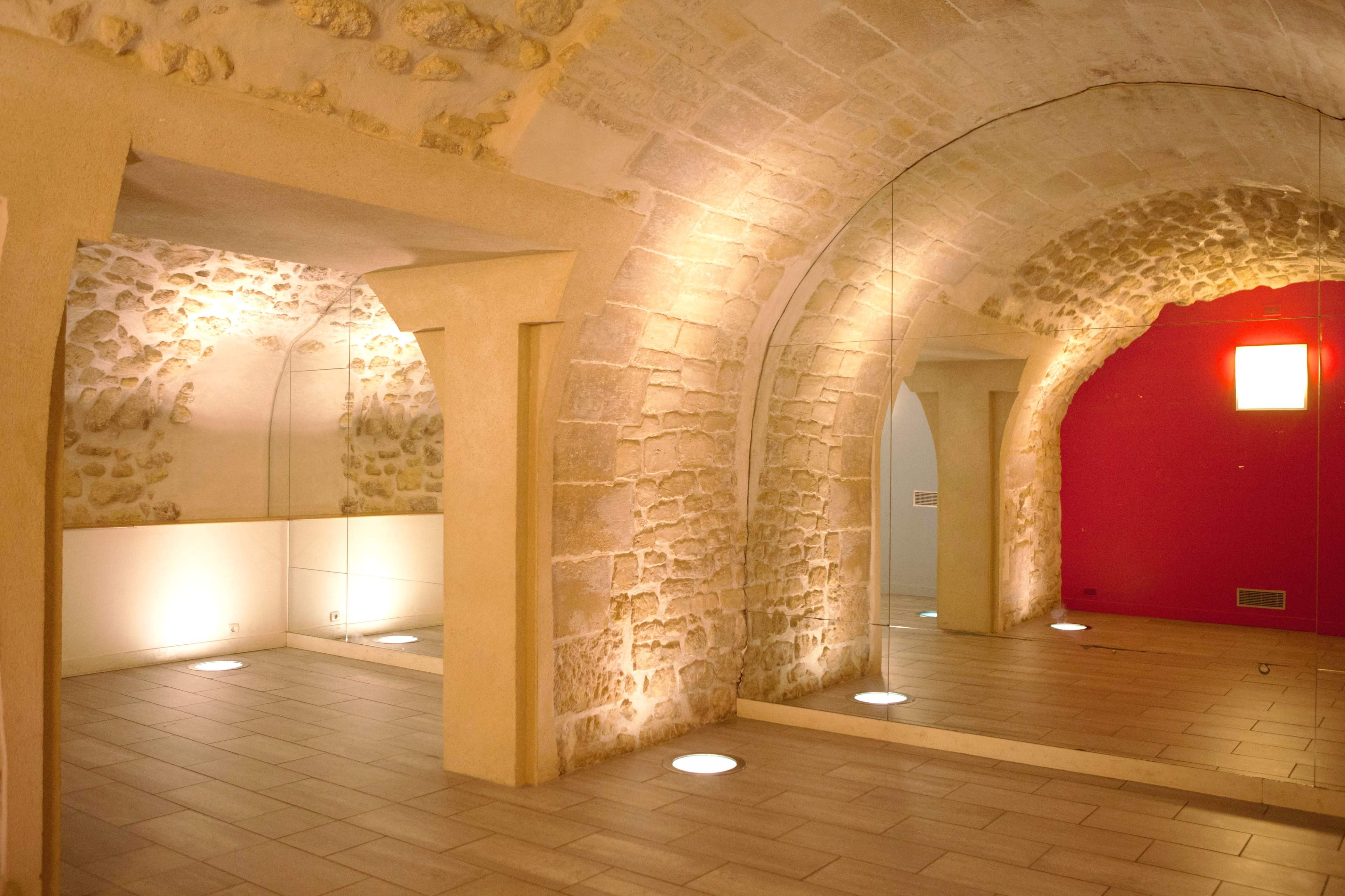 The image depicts a well-lit, stone-walled room with arched ceilings and a mirrored wall, contrasting with a vibrant red wall on one side.