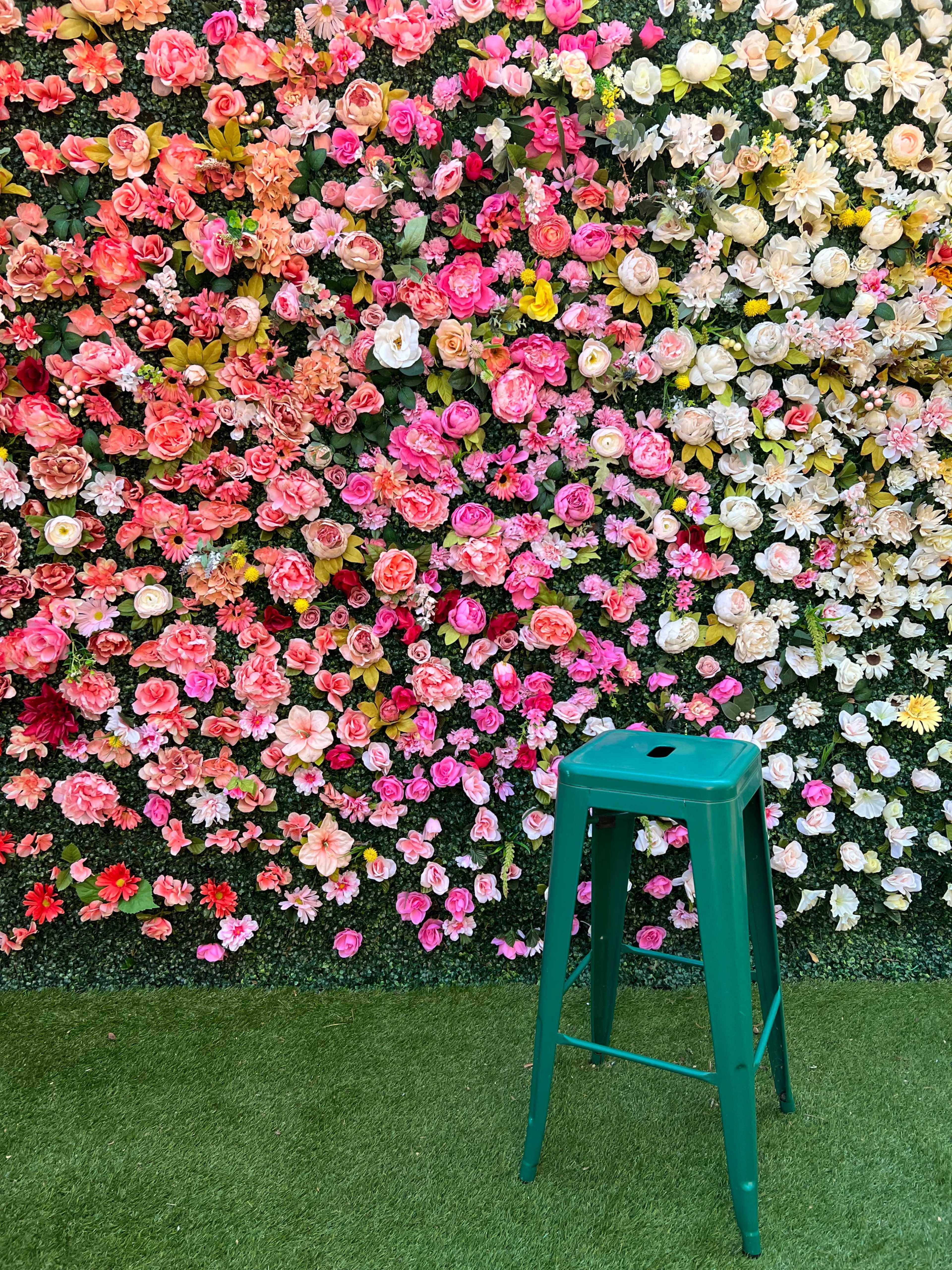 A green metal stool stands in front of a vibrant floral backdrop filled with various colorful flowers.