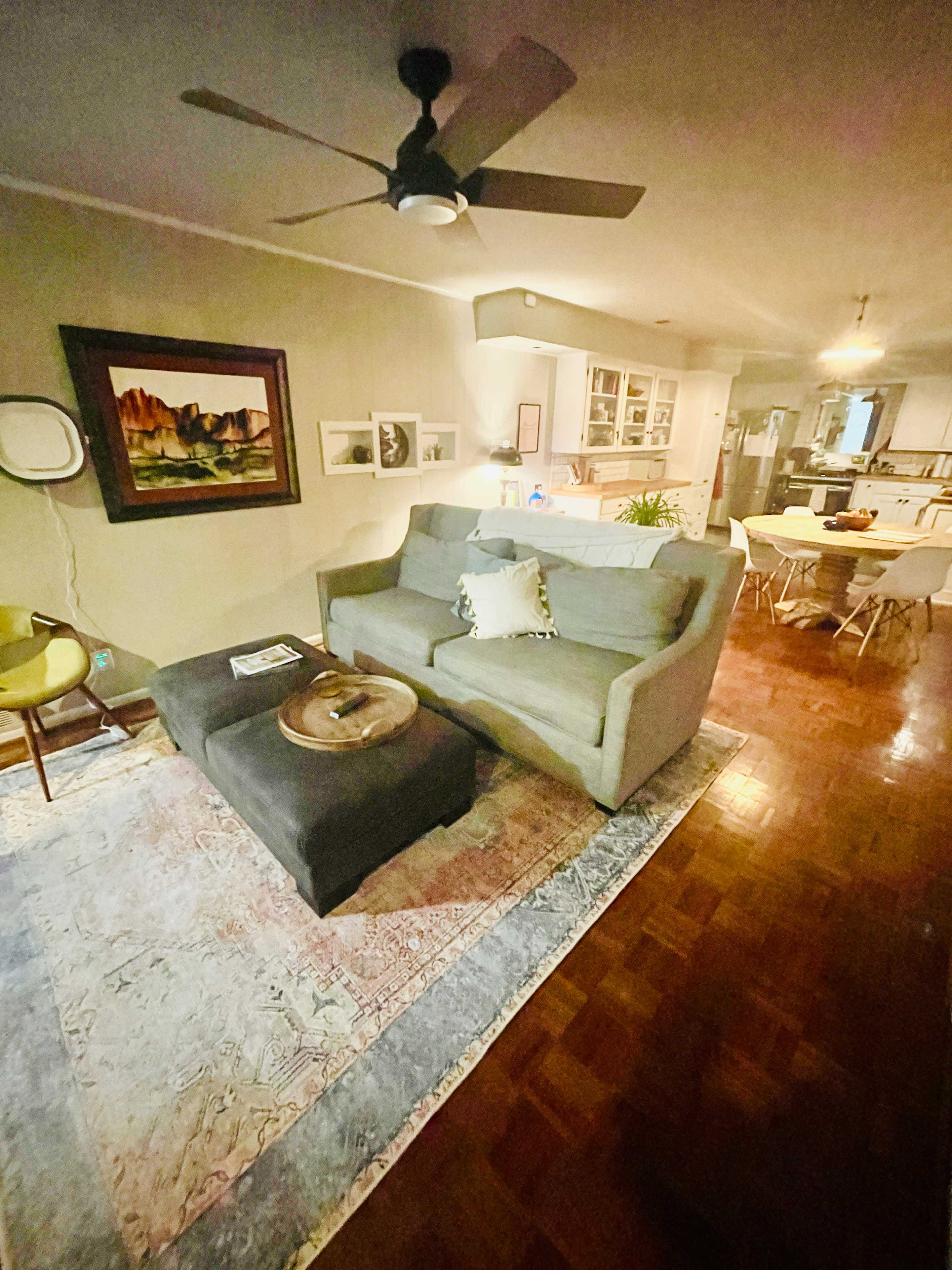 The image shows a cozy living room with a gray sectional sofa, a round coffee table, a patterned area rug, and a dining area visible in the background.