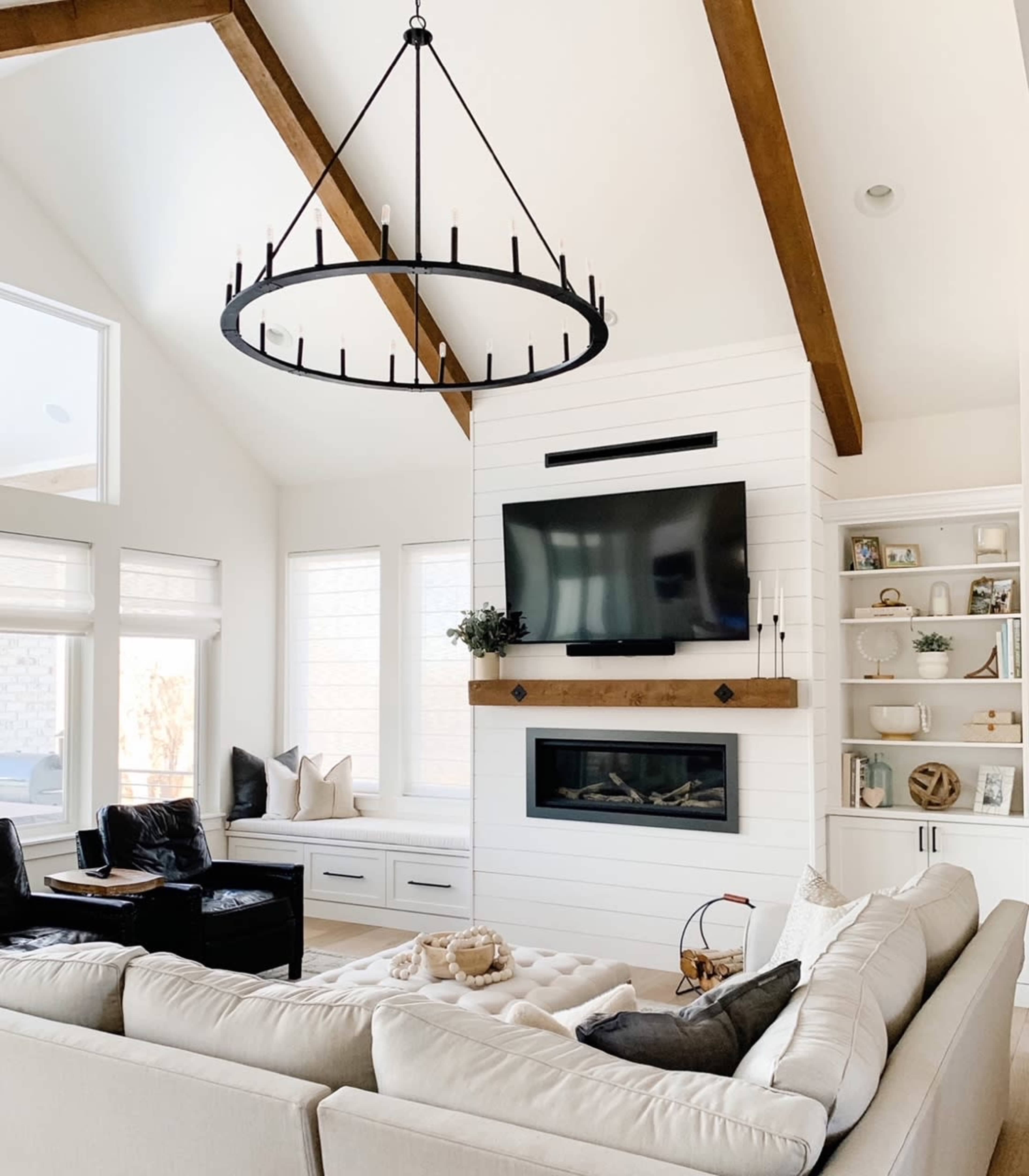 The photo shows a modern living room with a large circular chandelier, a flat-screen TV mounted above a wooden mantel, and a sectional sofa facing a fireplace.