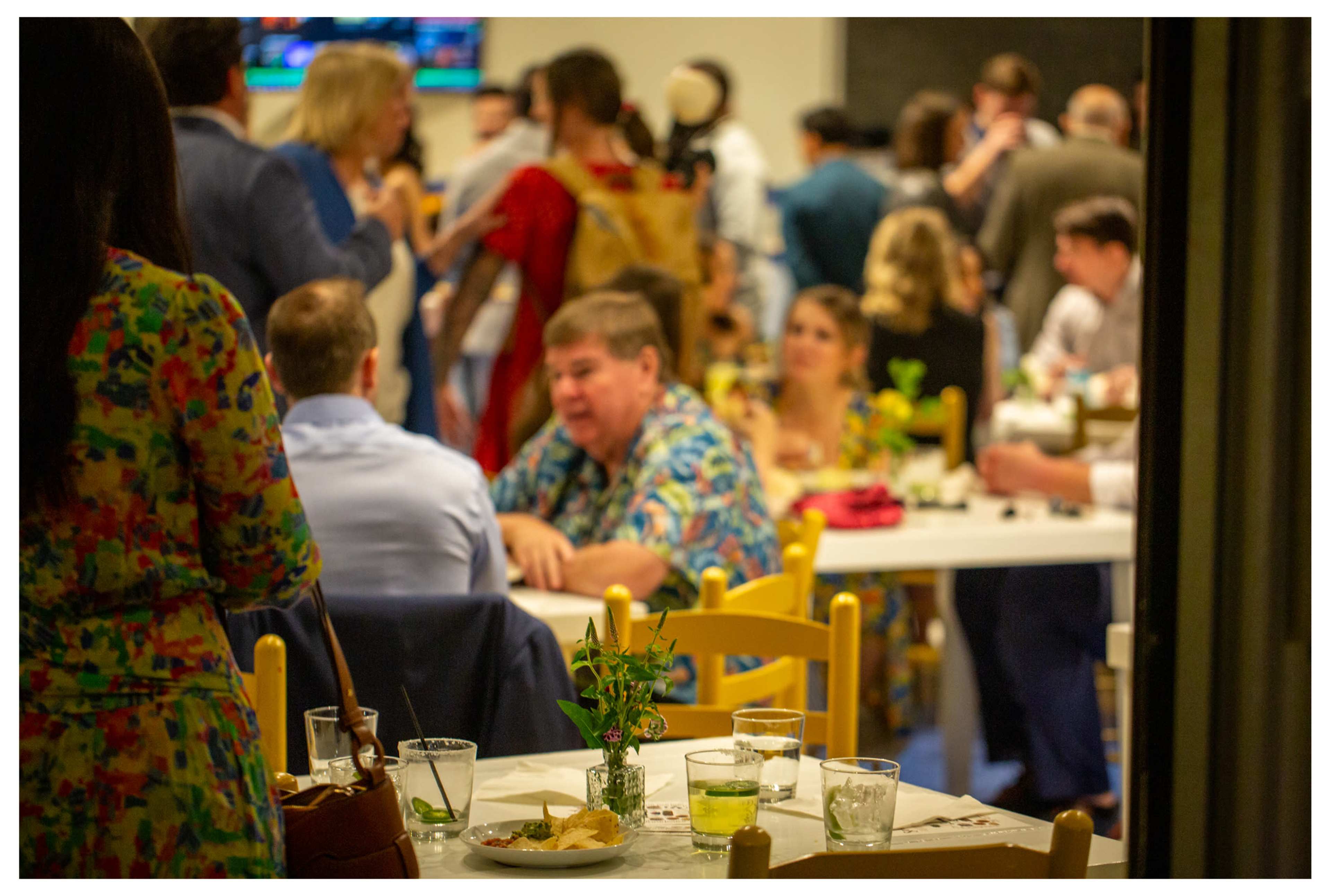 A crowded restaurant scene features people socializing at tables, with drinks and food in the foreground.