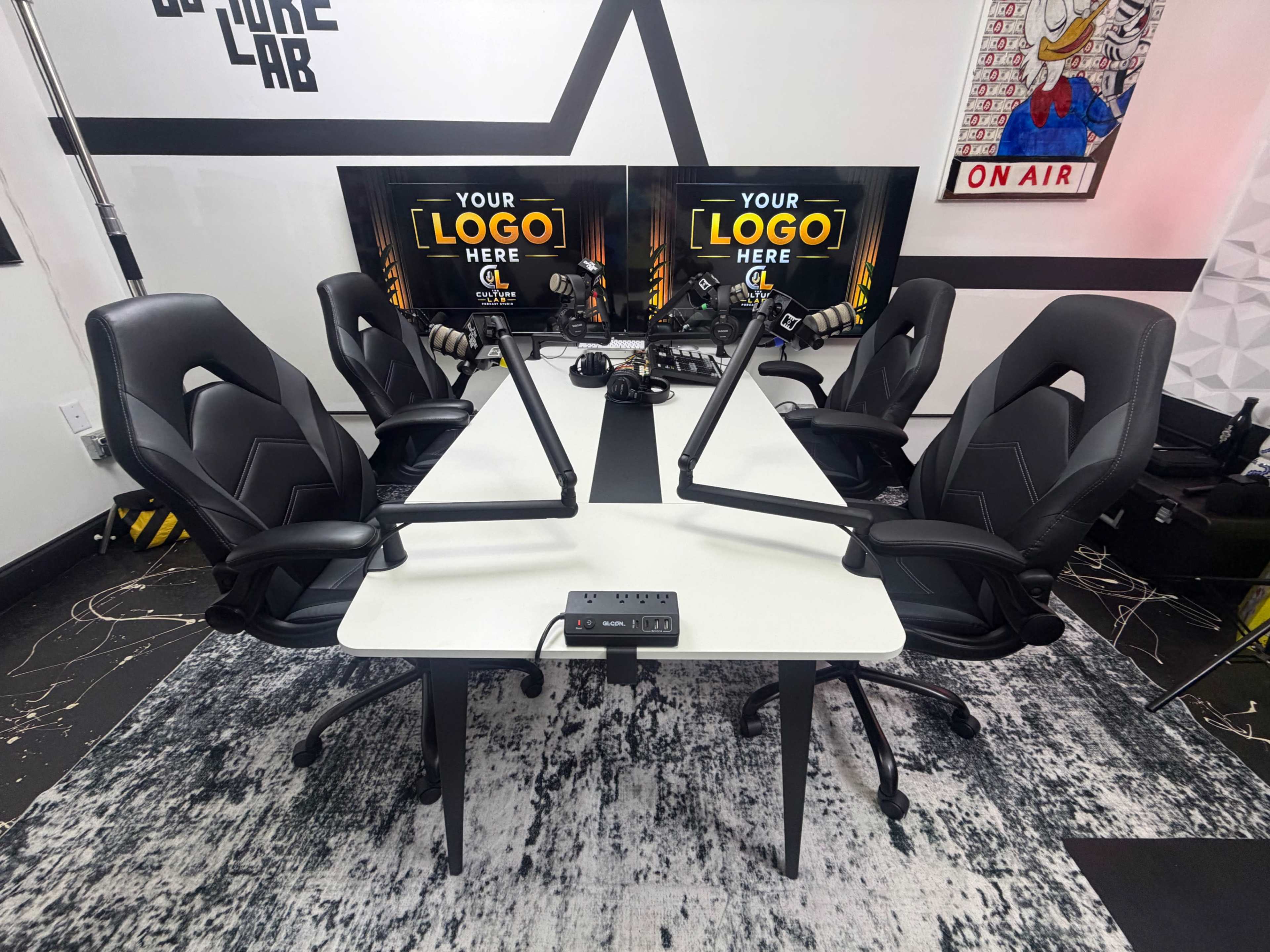 The image shows a modern podcasting studio with two desks equipped with adjustable microphone arms and black ergonomic chairs, alongside two monitors displaying a placeholder for a logo.
