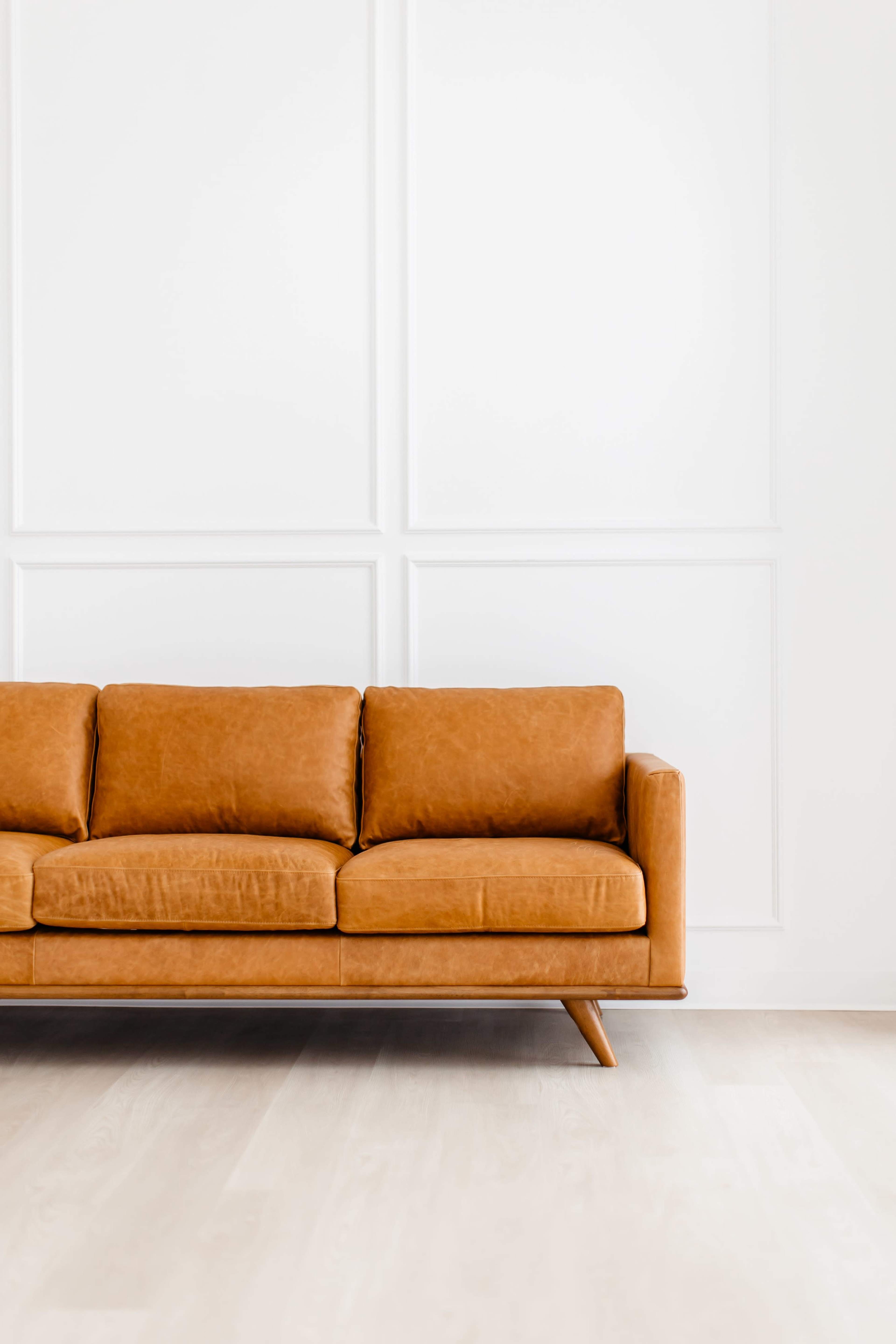 A brown leather sofa with wooden legs is placed against a white wall with paneling.