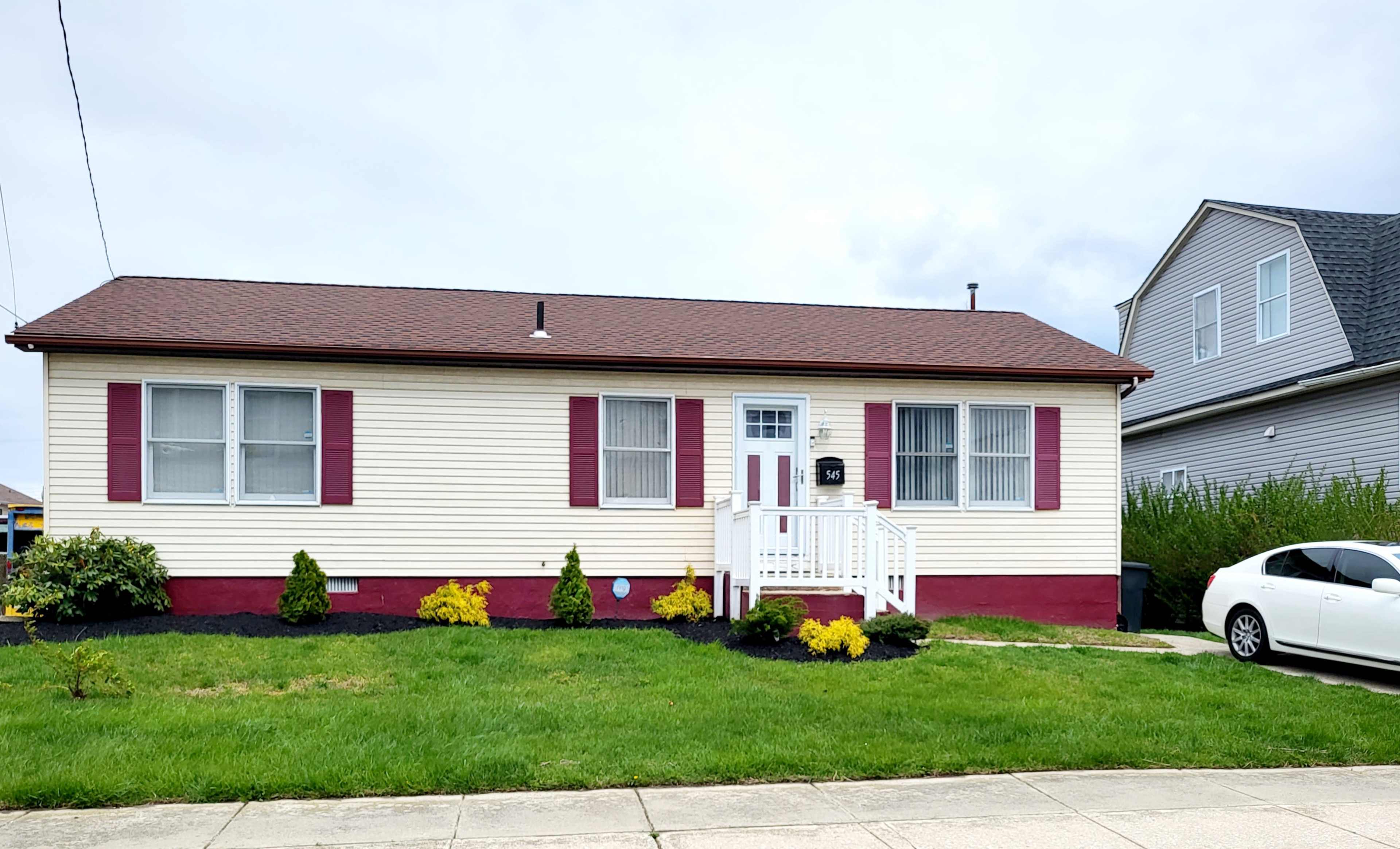 A single-story house with beige siding, red shutters, and a small front porch, surrounded by a well-maintained lawn and landscaped shrubs.