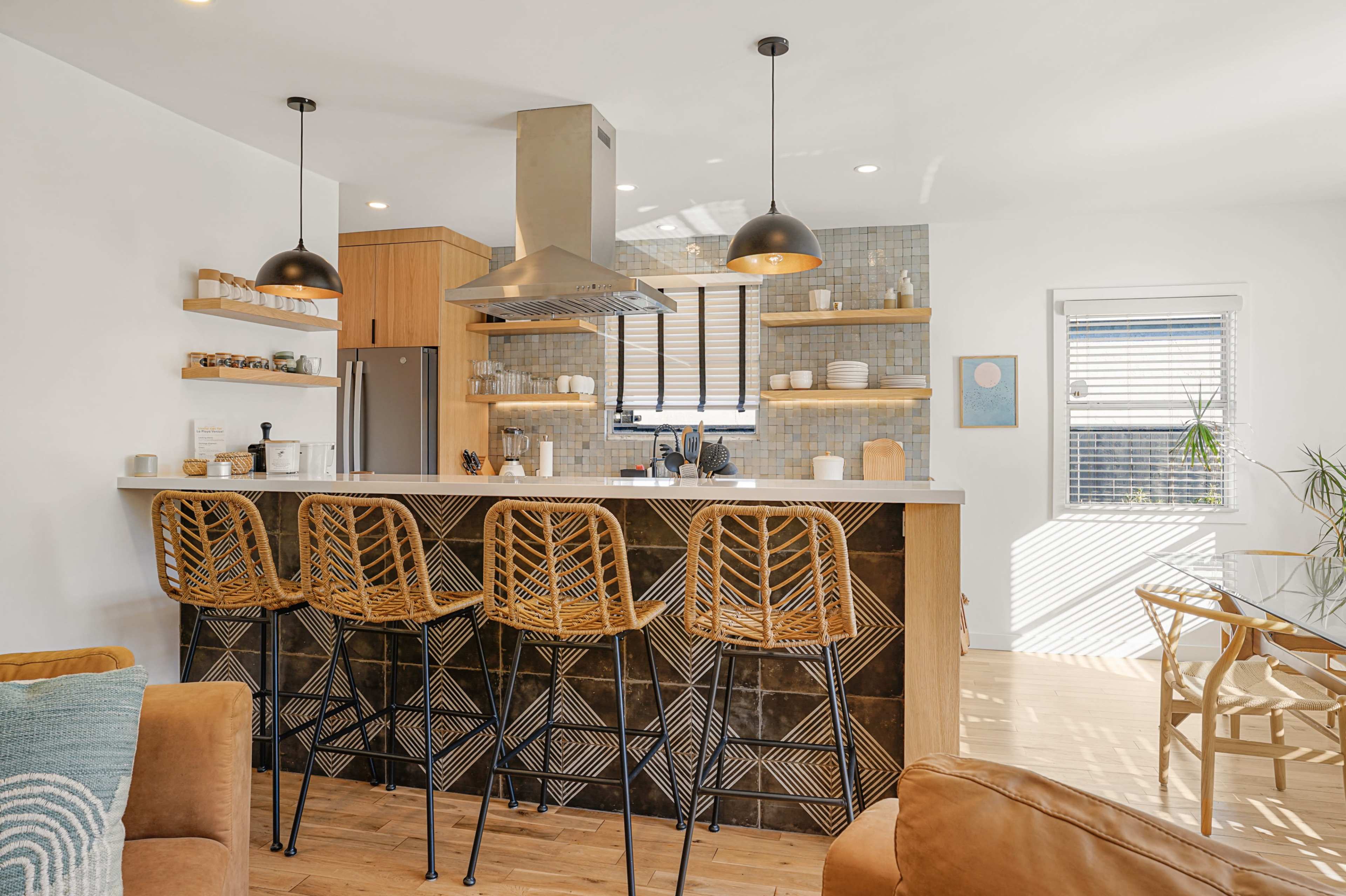 A modern kitchen features a counter with four rattan bar stools, stainless steel appliances, and a decorative tile backsplash.