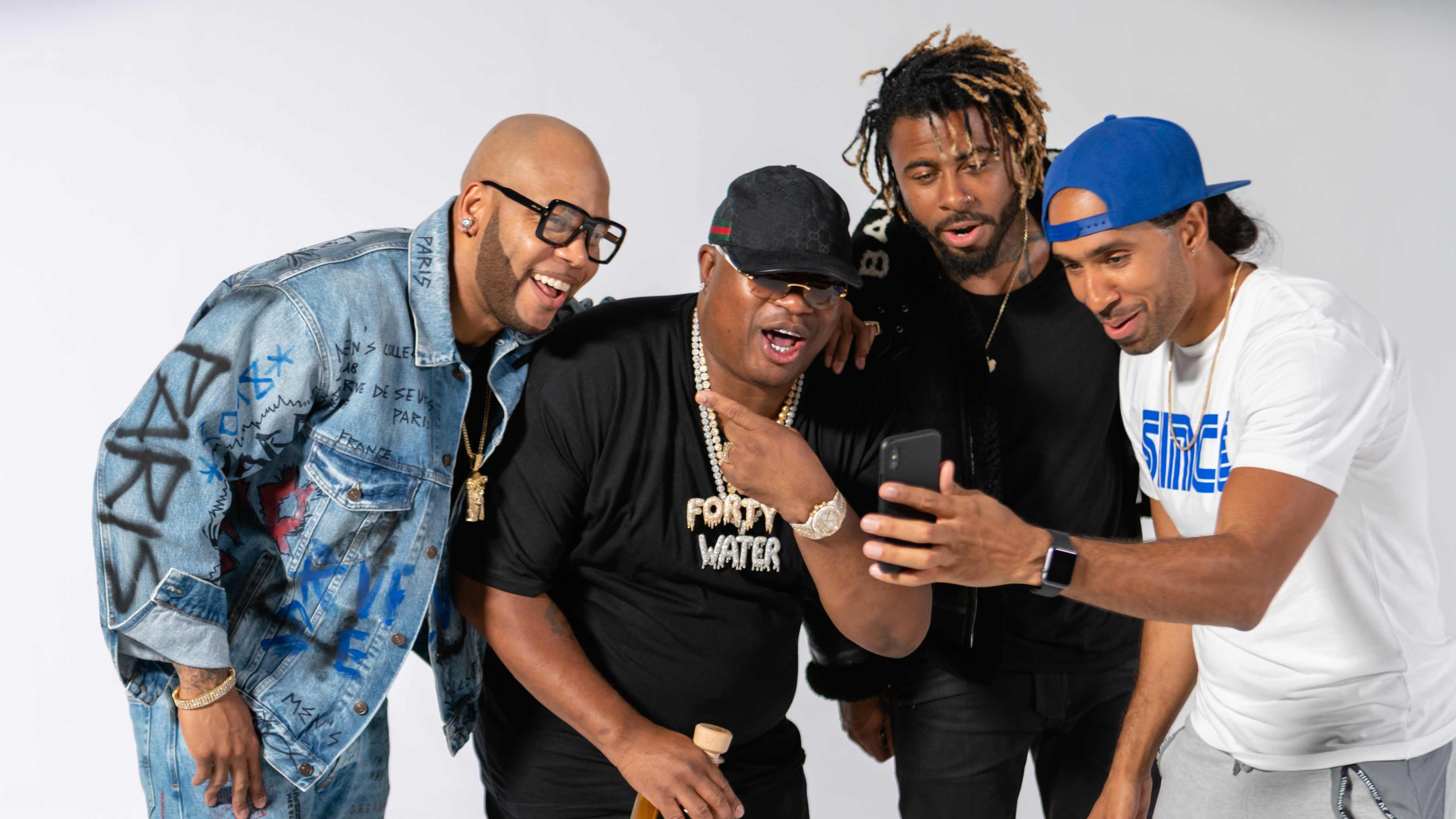 Four men are gathered together, smiling and looking at a phone in a well-lit, neutral background.