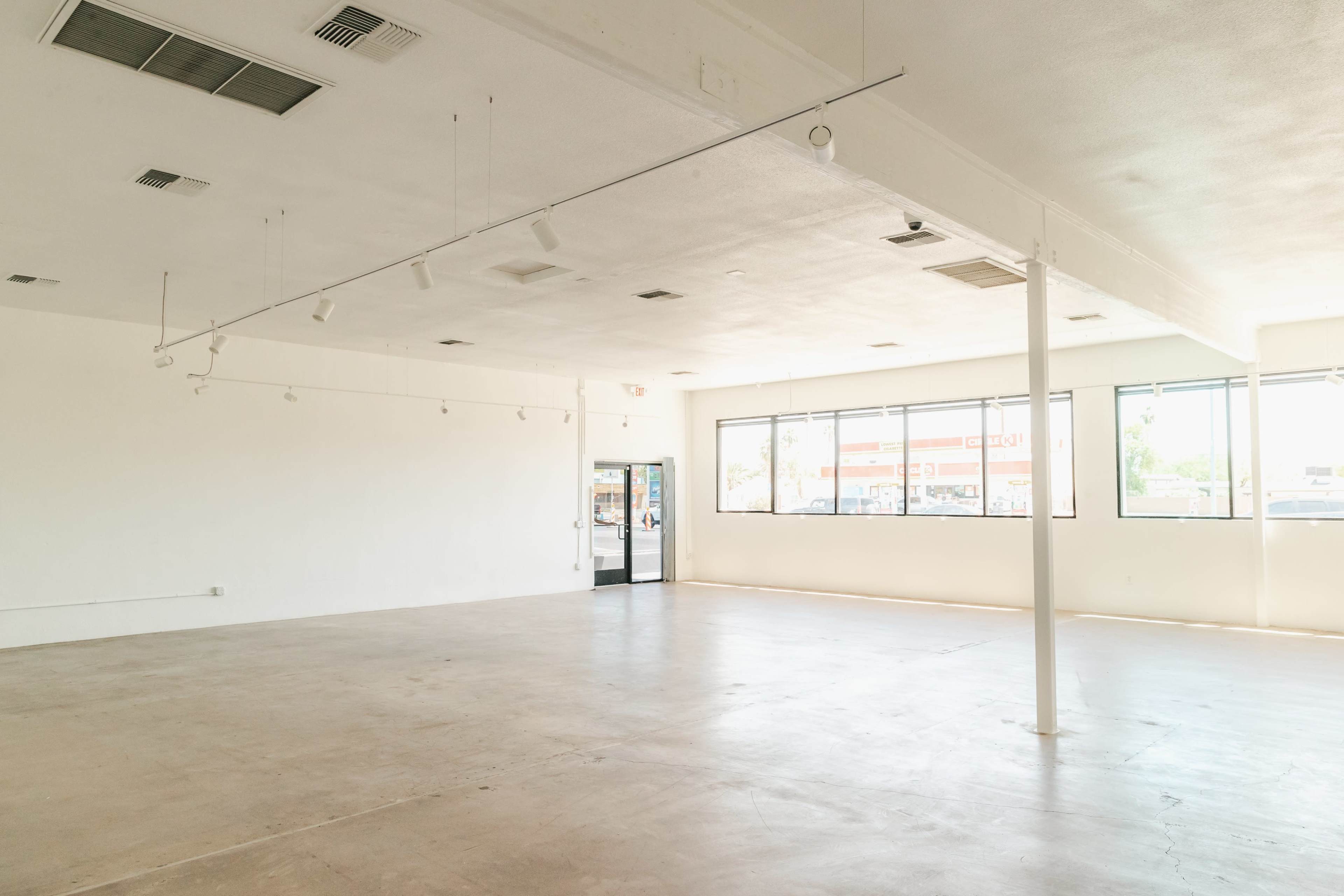 The image shows an empty, well-lit interior space with white walls and a concrete floor, featuring large windows and a single support column.