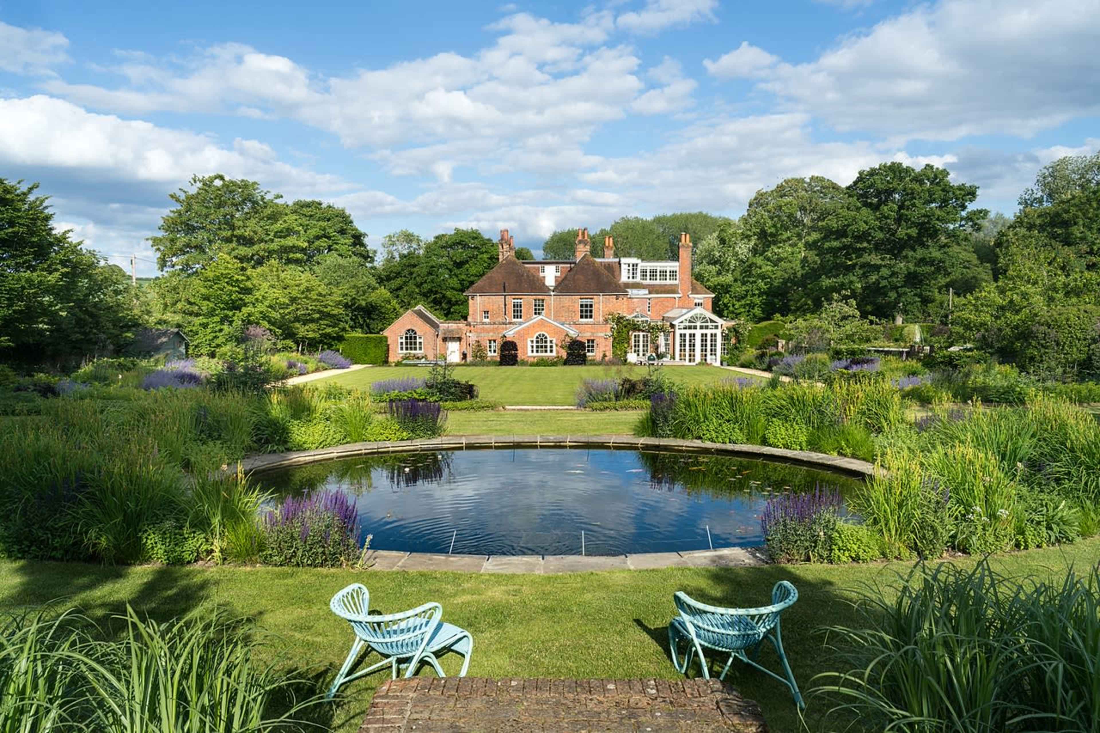 A large red-brick house is surrounded by a well-maintained garden with a circular pond and green chairs in the foreground.
