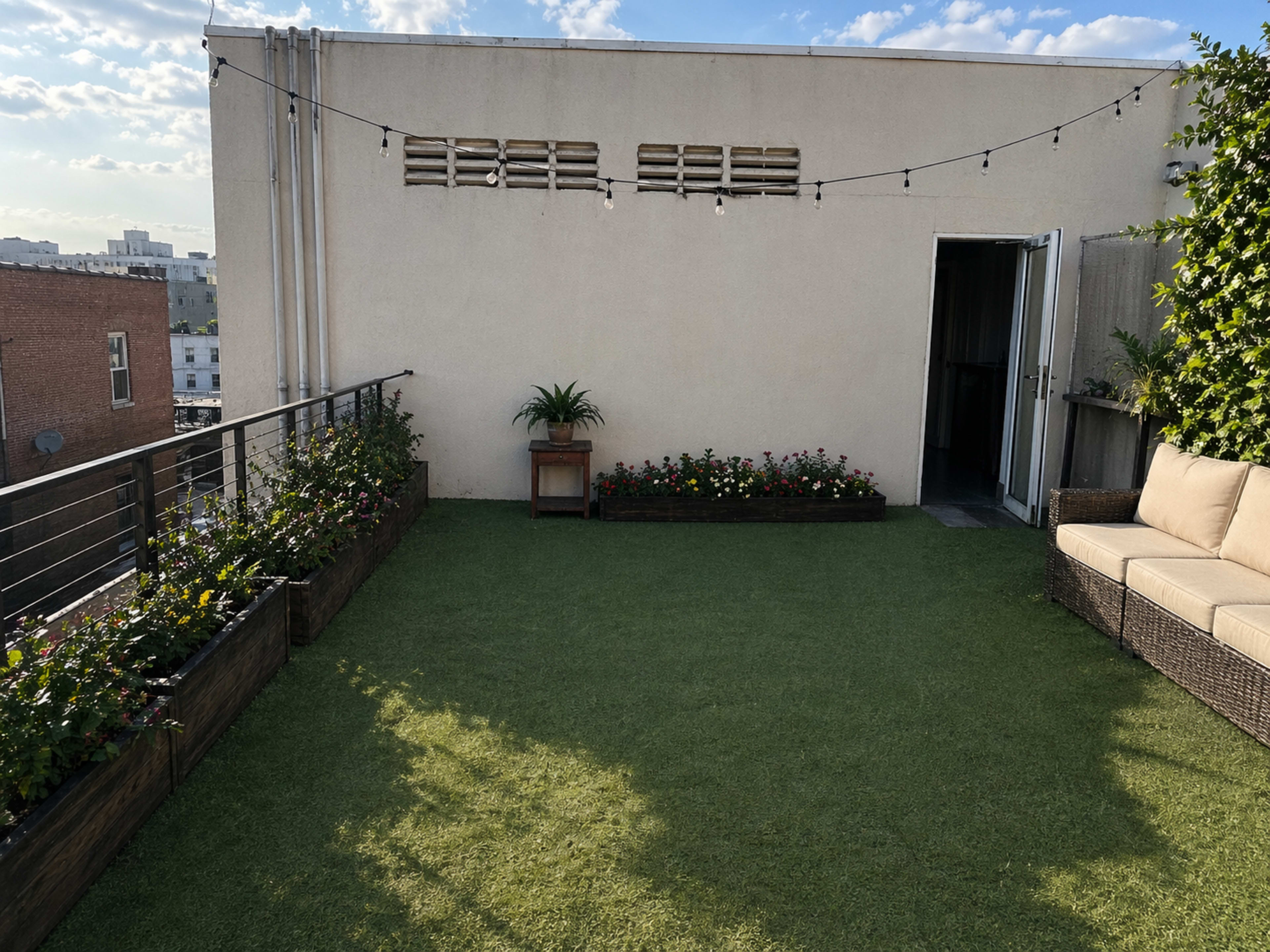 The image shows a rooftop terrace with artificial grass, planters filled with flowers, a potted plant, a seating area, and a doorway leading inside.