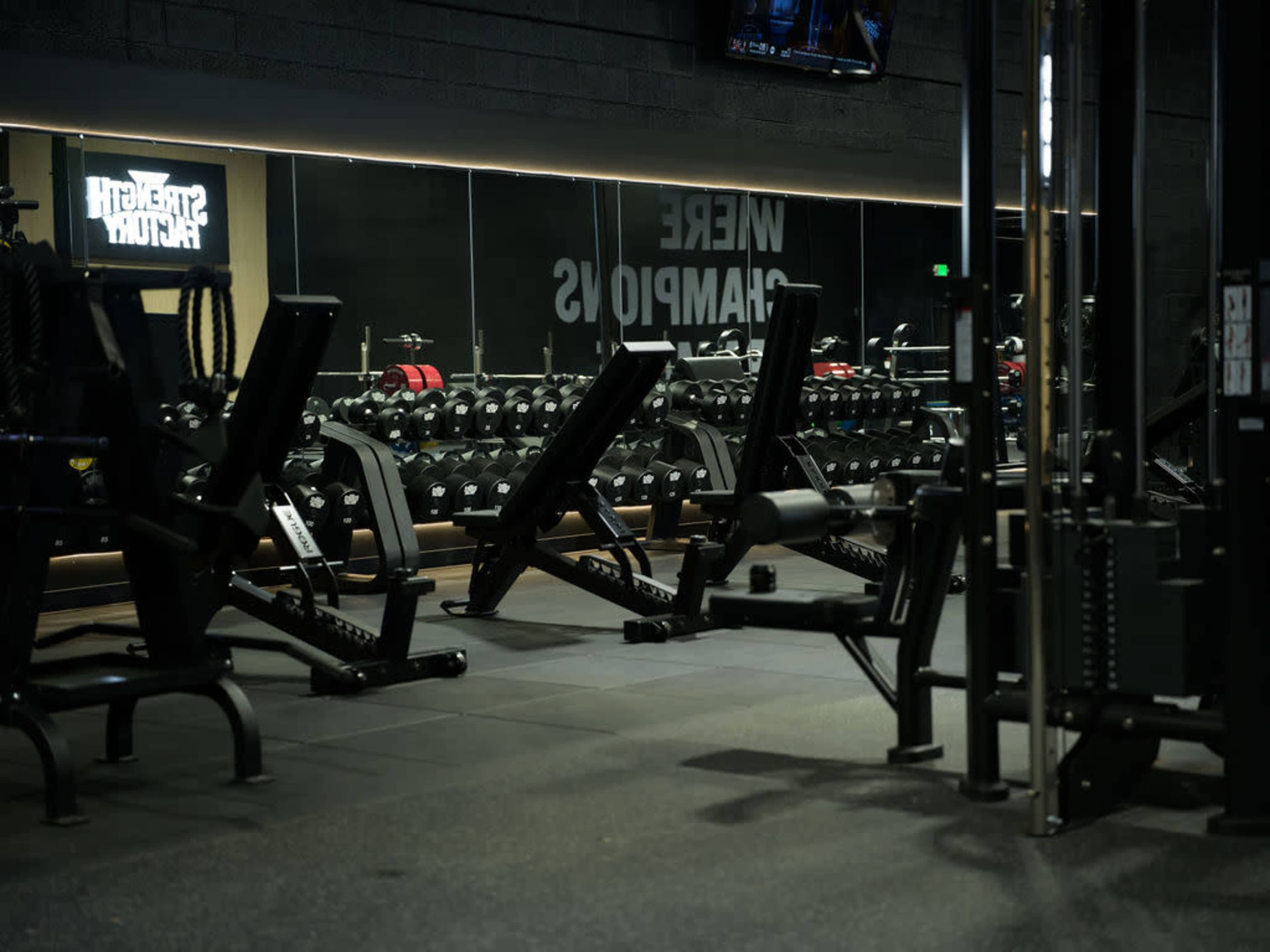The image shows an indoor gym with rows of weightlifting equipment and free weights arranged neatly against a mirrored wall.