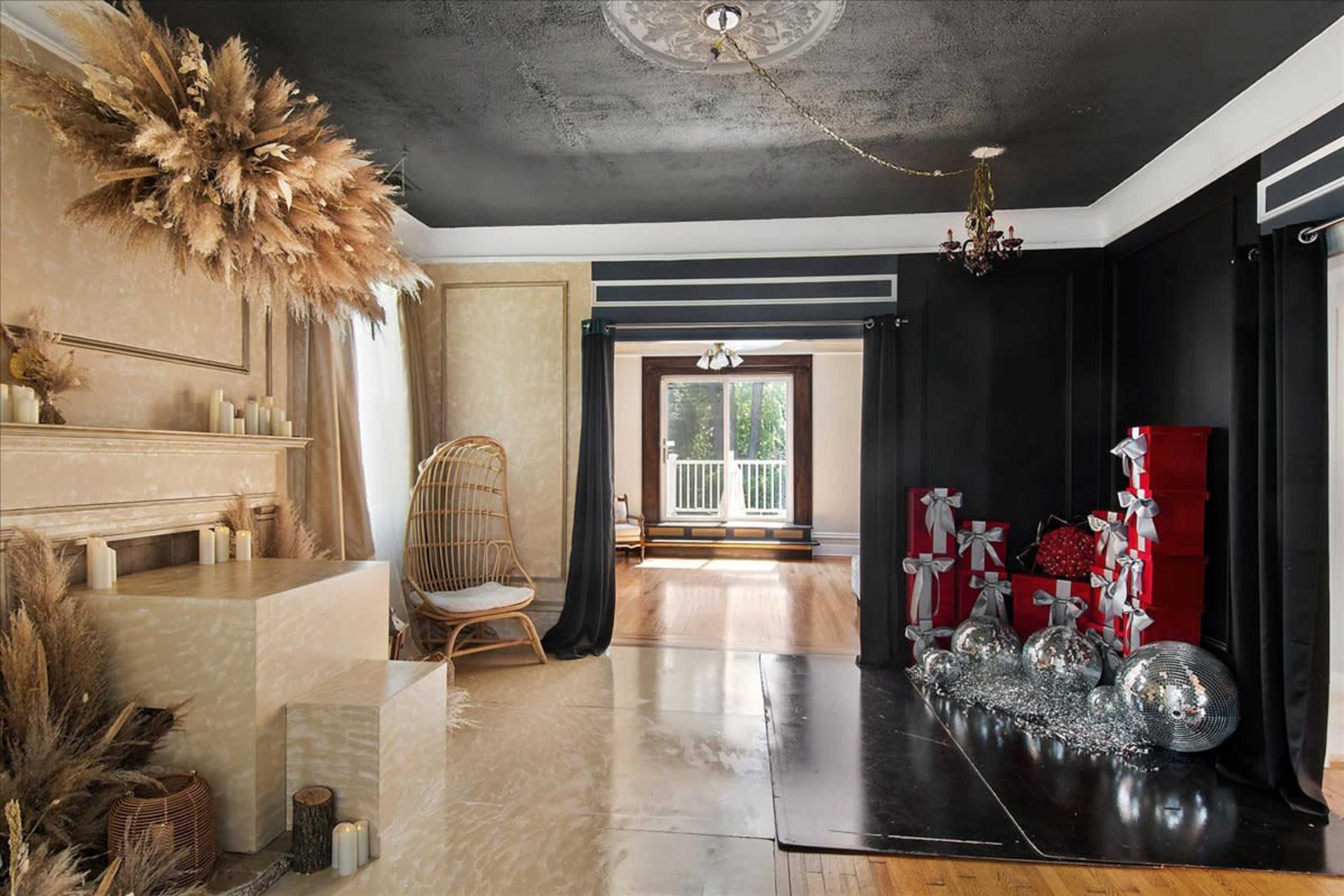 The image shows a stylish room featuring a decorative arrangement of pampas grass, candles, and wrapped gifts, along with a cozy seating area and large windows leading to a balcony.