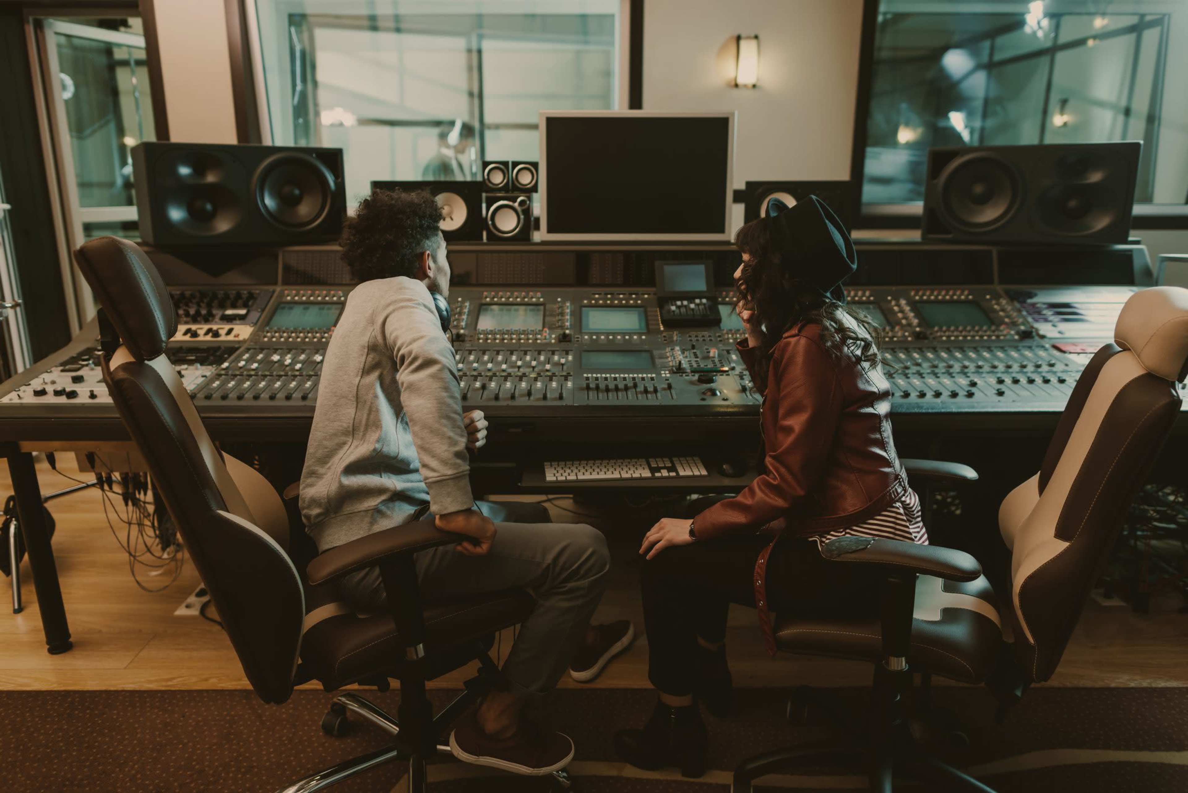 Two individuals are seated in front of a large music mixing console in a recording studio.