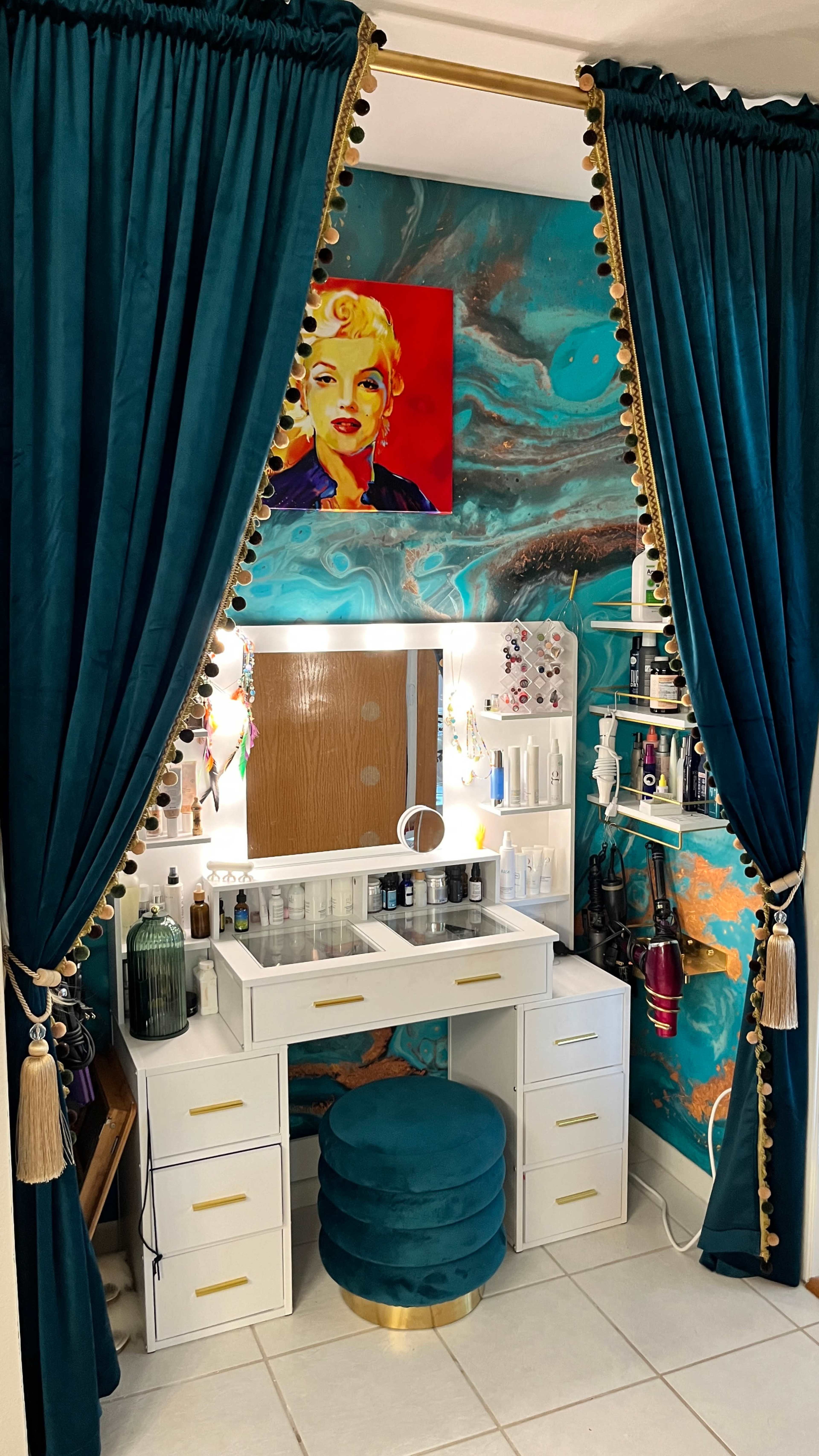 The image shows a vanity area with a white makeup desk, a round teal stool, and teal drapes adorned with decorative tassels, set against a colorful marbled wall with a portrait of a woman.