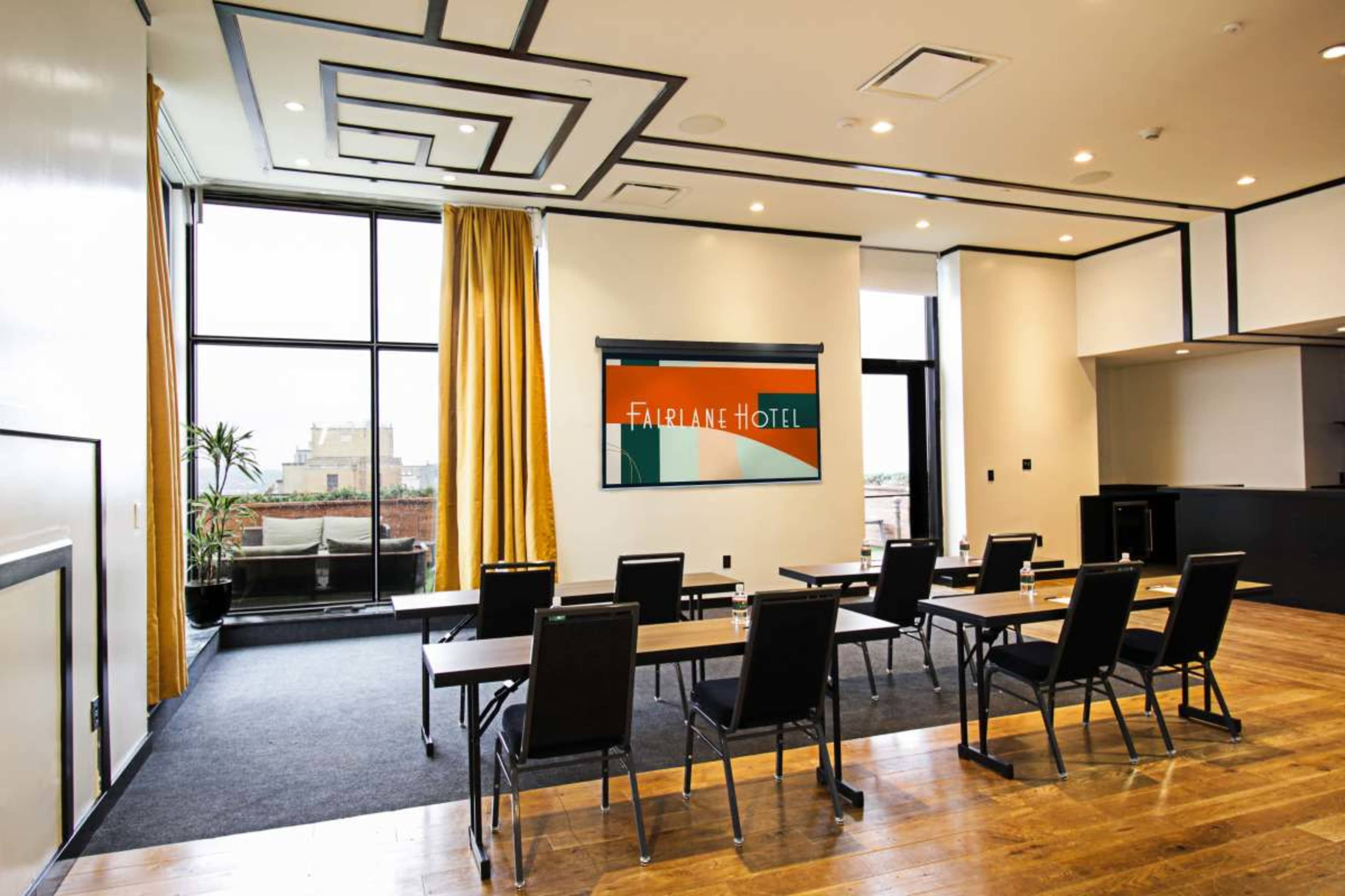 A conference room in the Fairlane Hotel features rows of tables and chairs, large windows with drapes, and a wall display showcasing the hotel’s logo.