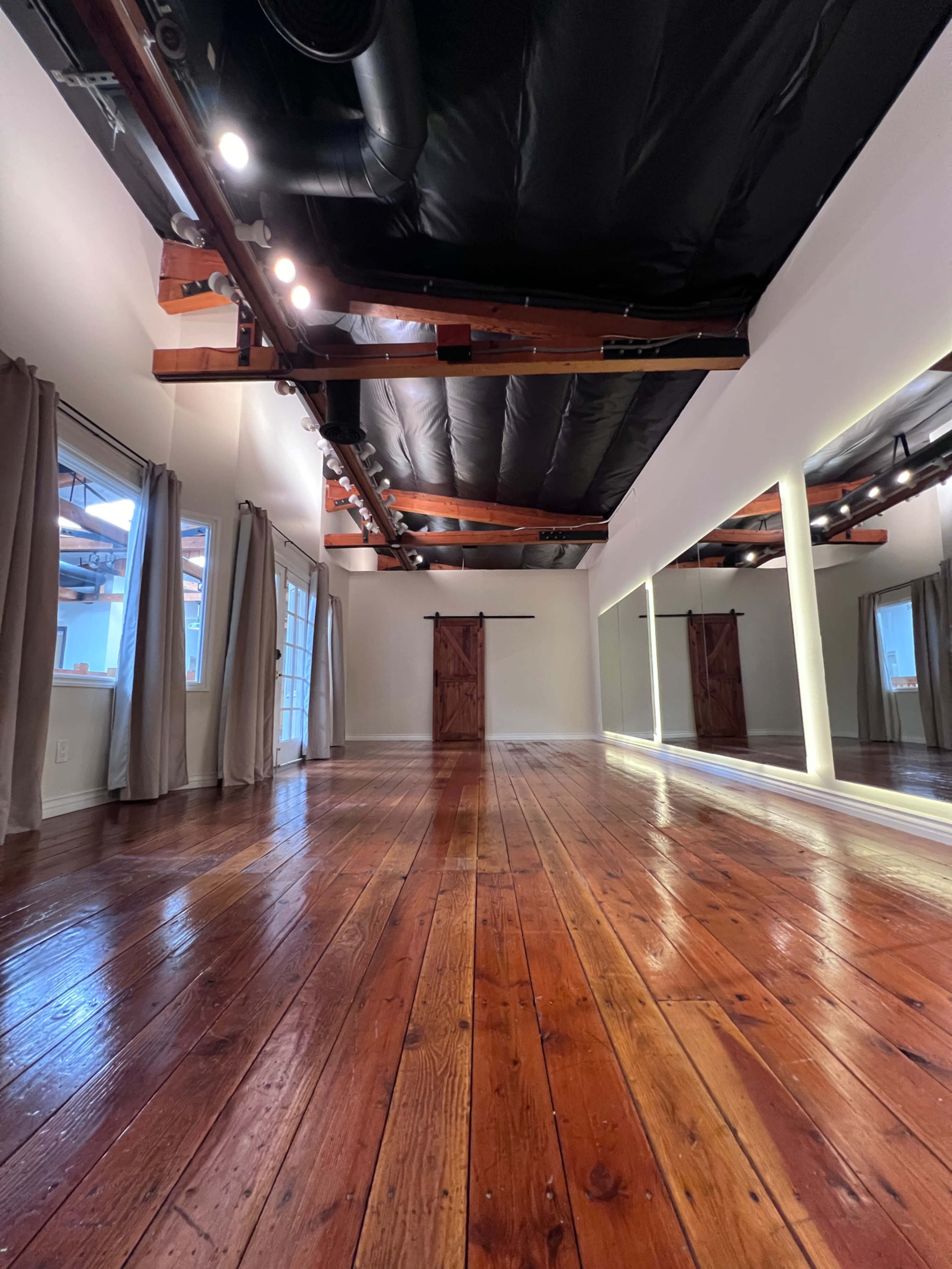 A spacious room features polished wooden floors, large windows with curtains, and a mirrored wall.