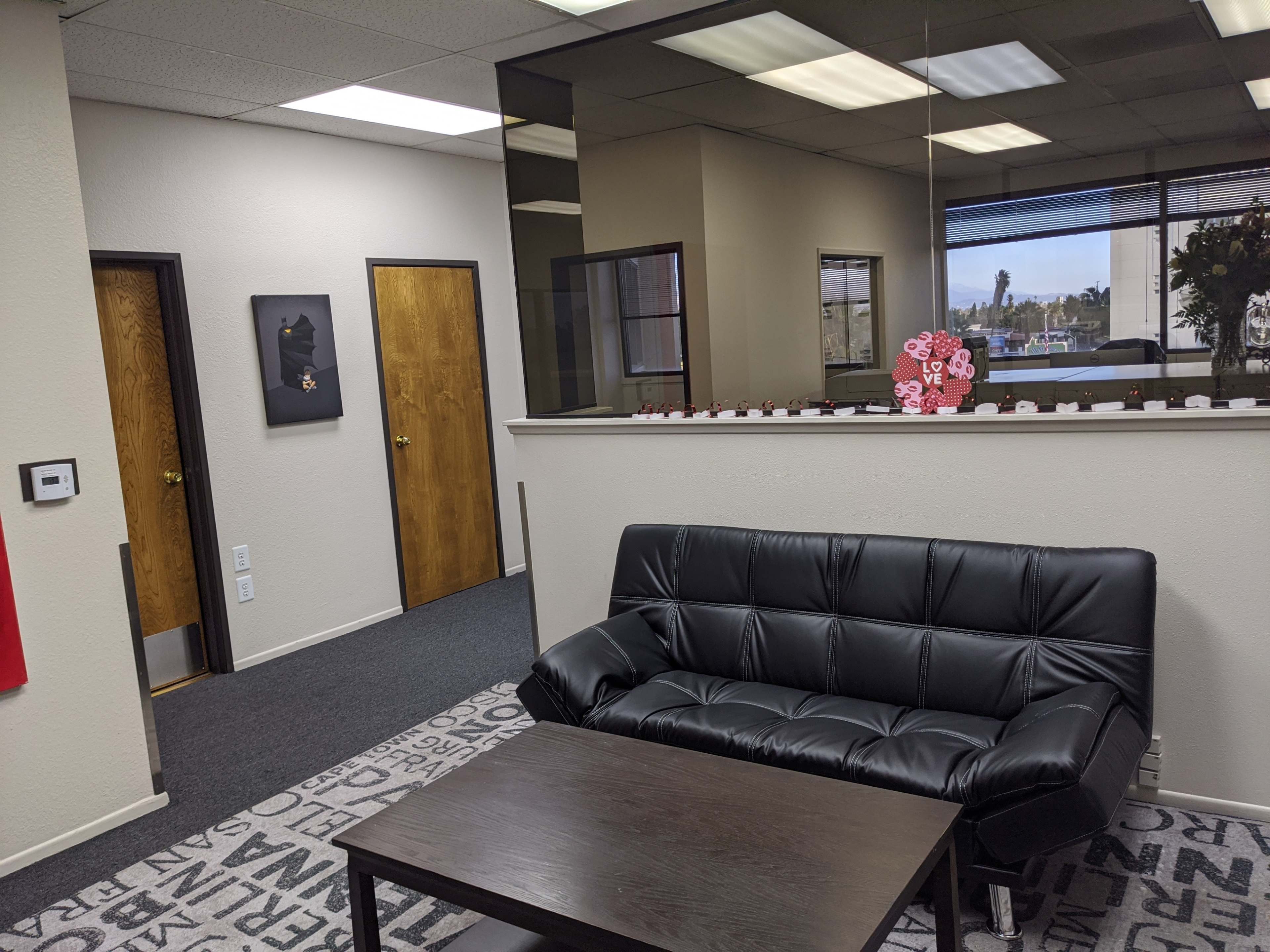 A modern office reception area with a black sofa and a small table, featuring two doors in the background and a reflective wall separating it from another room.