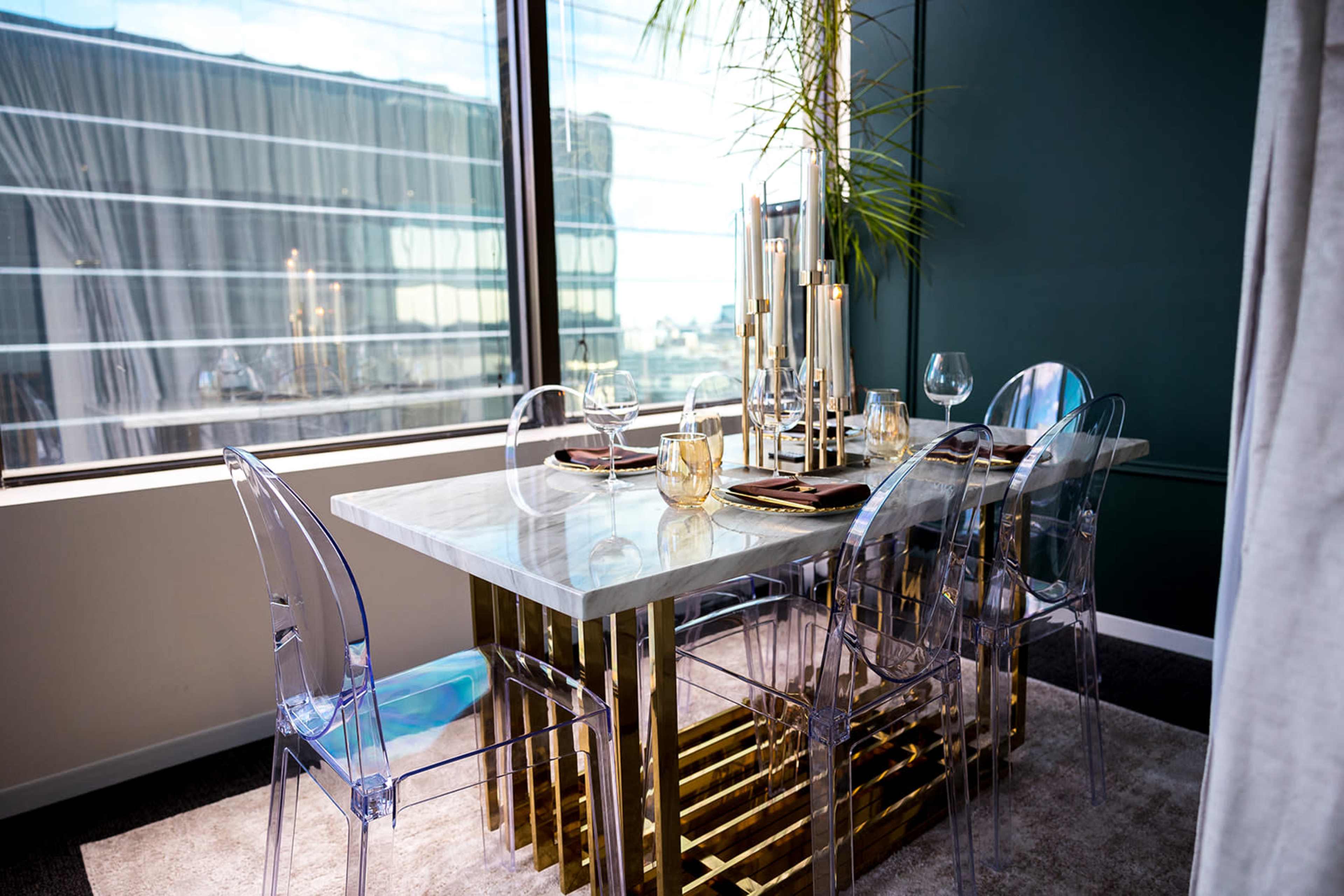 A modern dining area features a marble-topped table with gold accents, accompanied by transparent chairs and placed near a large window.