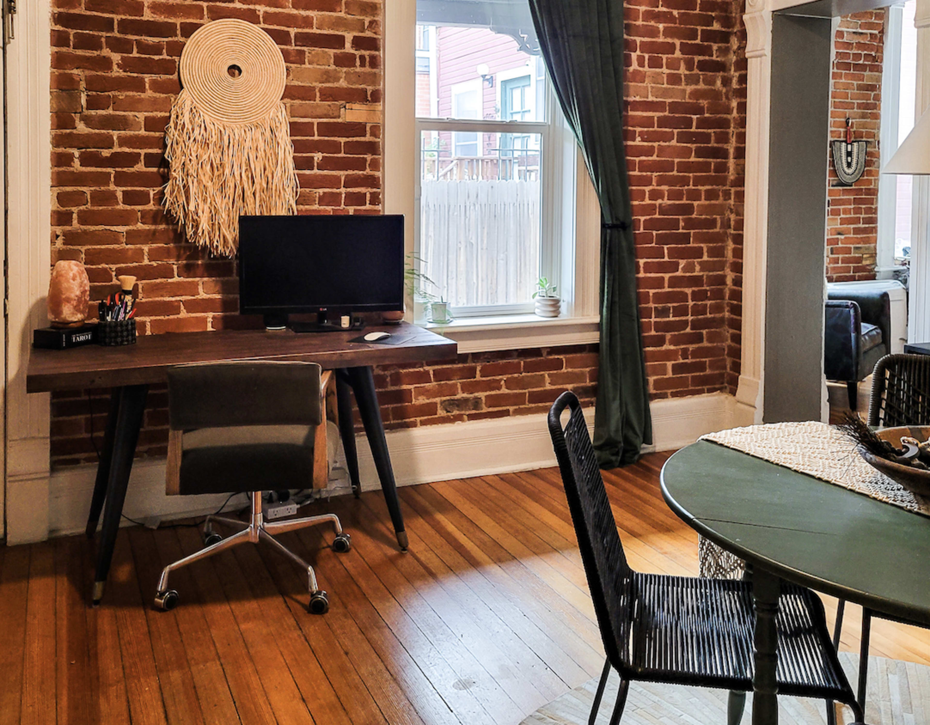 The image shows a room with exposed brick walls featuring a desk with a computer and a chair, alongside a round dining table and green curtains.