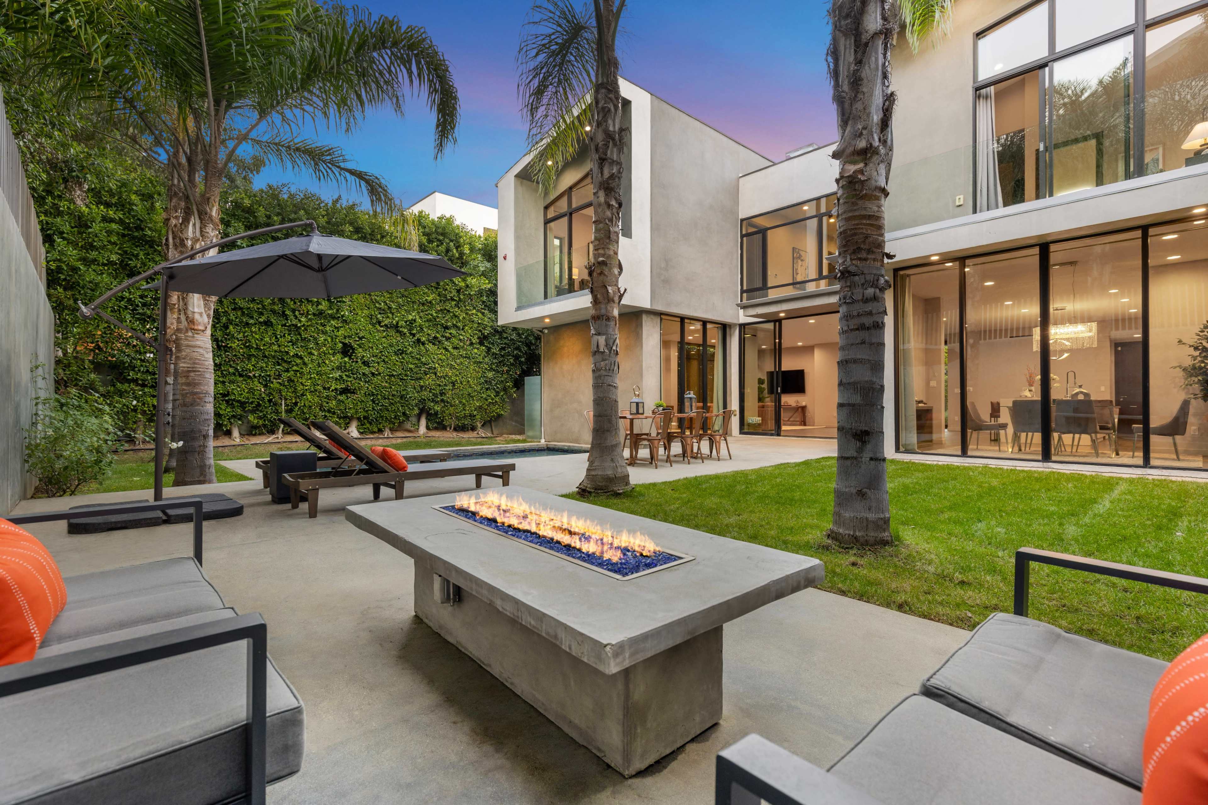 The image shows a modern outdoor seating area with a fire pit, surrounded by palm trees and a contemporary house with large glass windows.