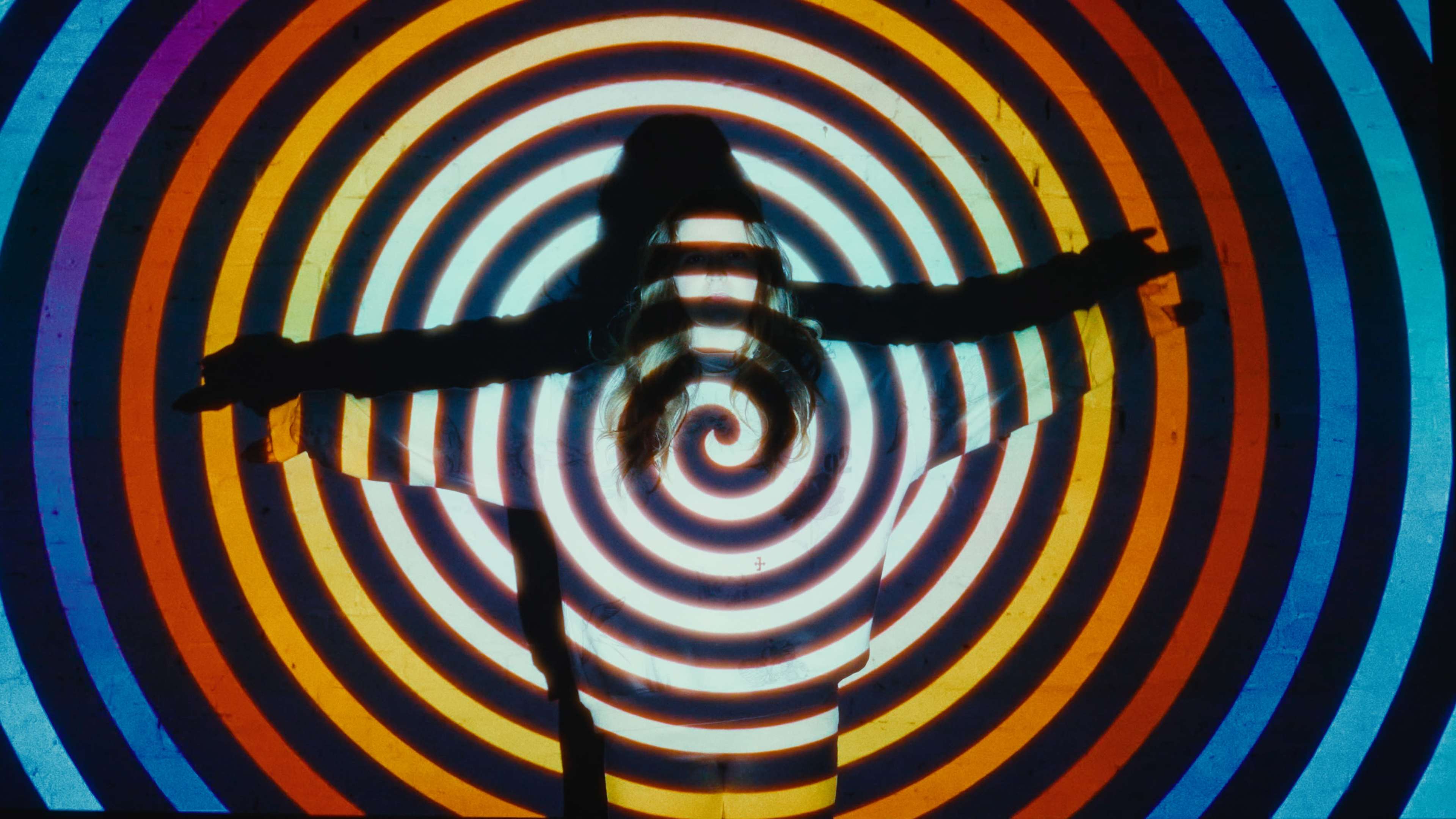 A person stands with arms outstretched in front of a brightly colored spiral pattern projected on a wall.