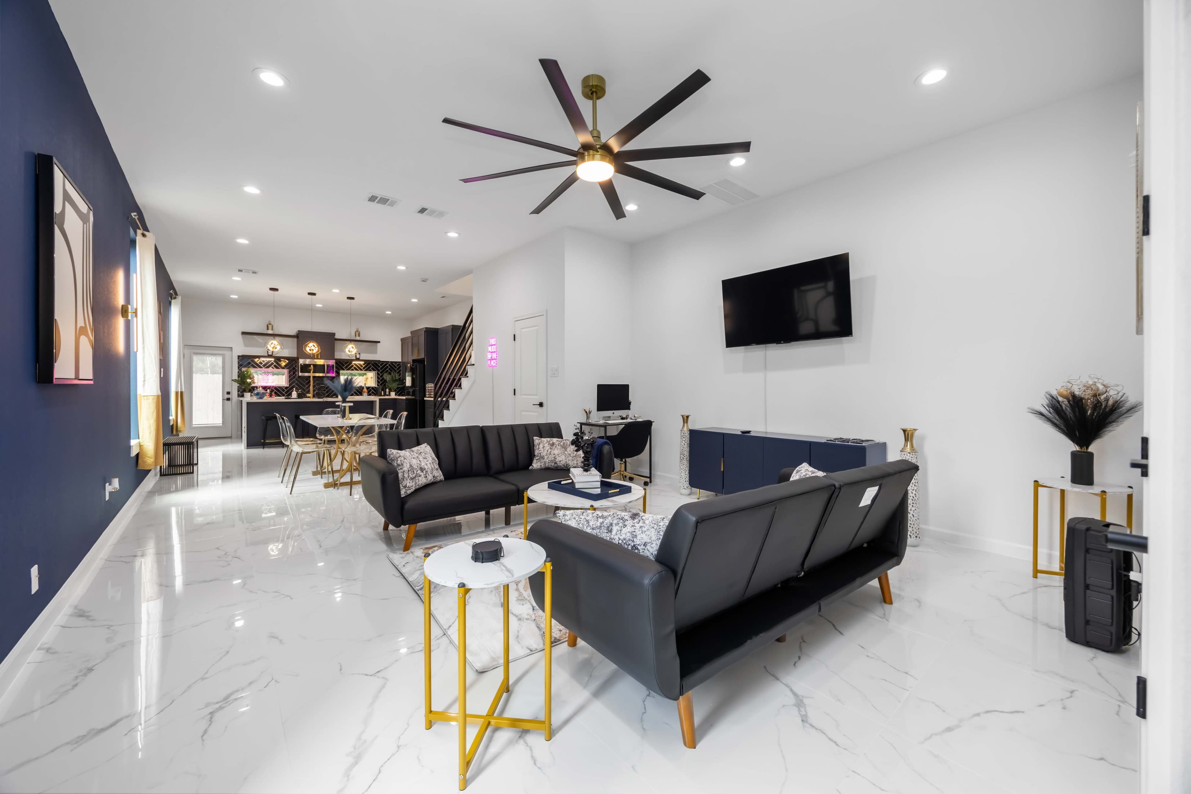 The image shows a modern living room with two black sofas, a large ceiling fan, and a television mounted on the wall.