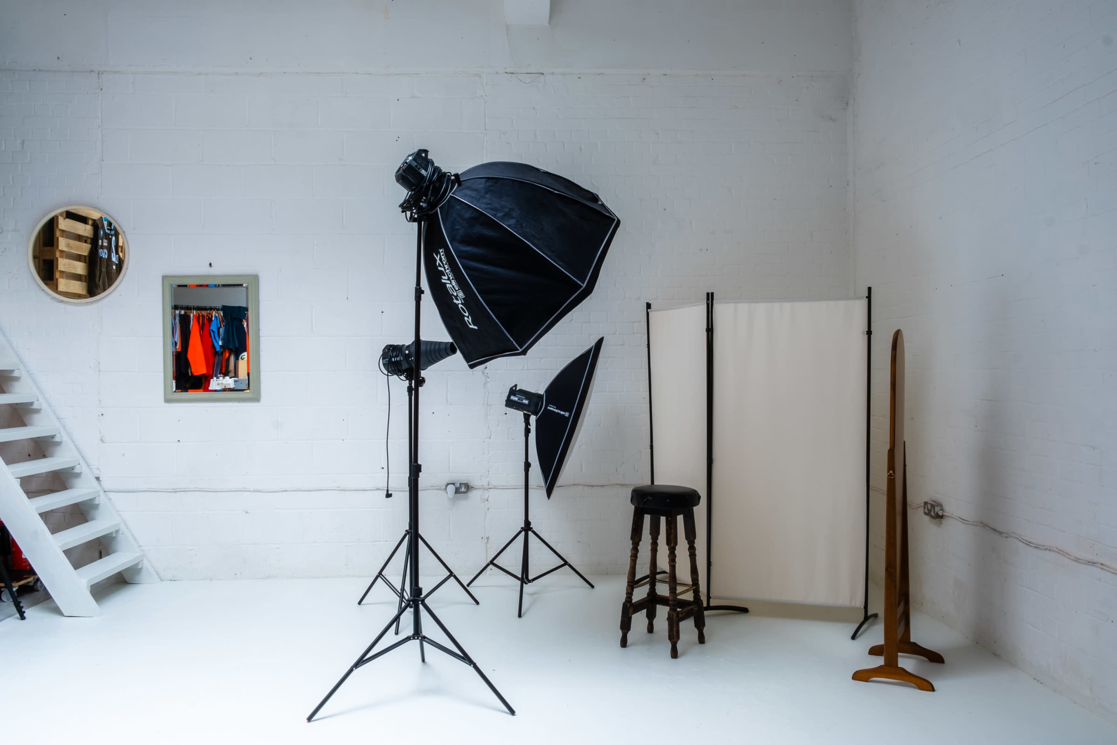 A photography studio with three softbox lights, a stool, a standing mirror, and a folding screen in a bright, white-walled space.